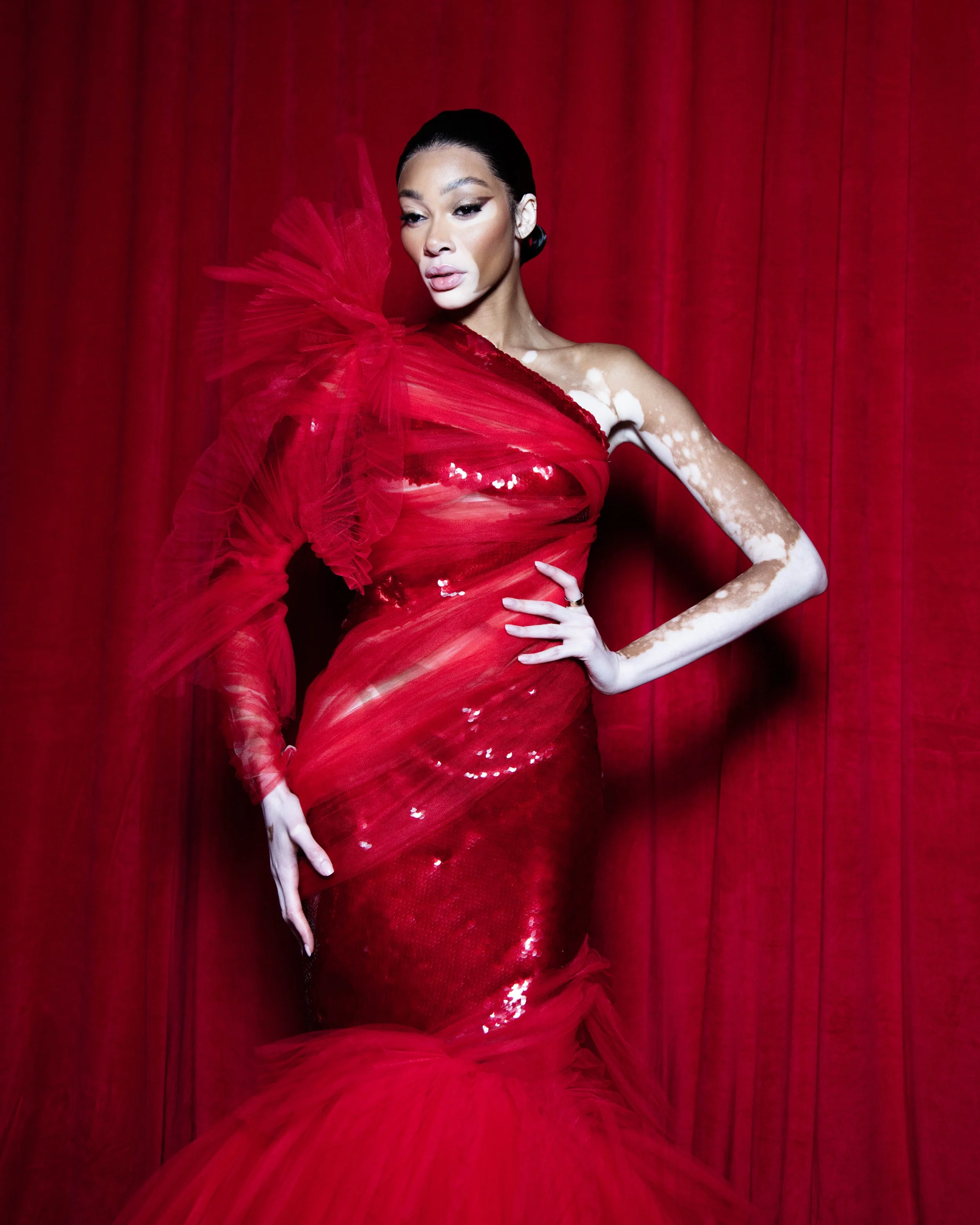 Woman in a red, form-fitting, sequin dress with tulle embellishments, standing in front of a red curtain.