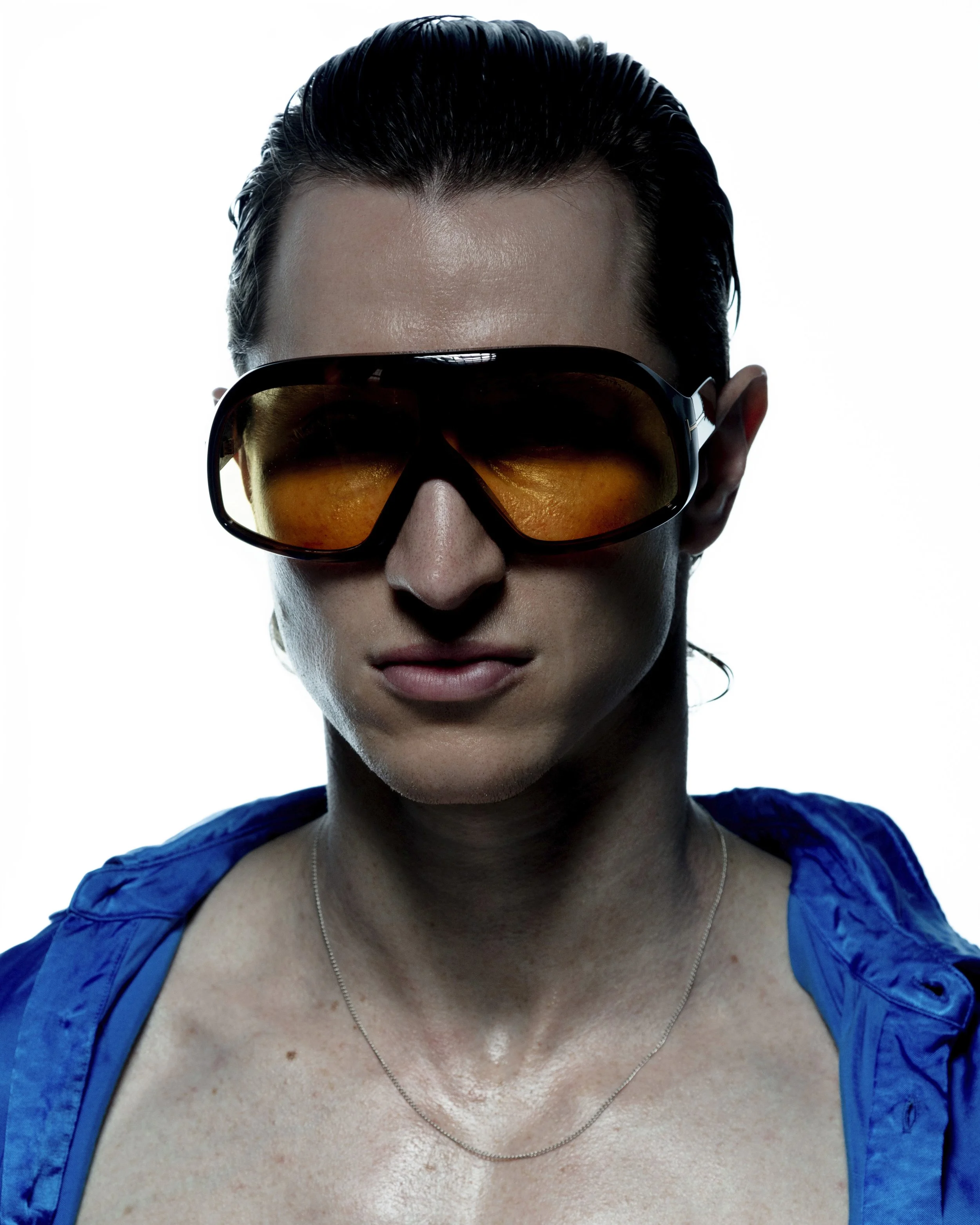 A person wearing large orange-tinted sunglasses, a blue jacket, and a thin chain necklace, with slicked-back dark hair and a serious expression.