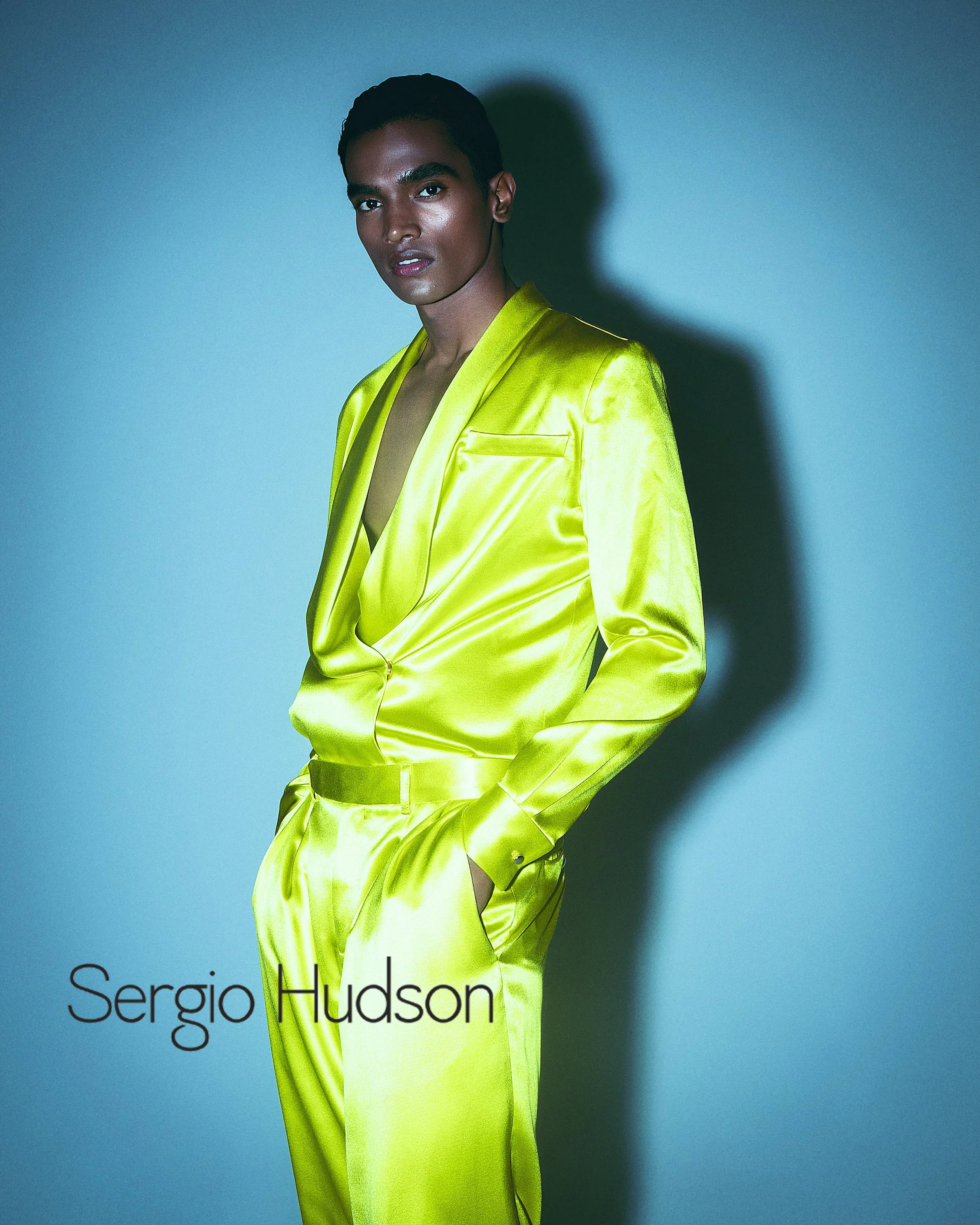 A man in a shiny, lime green satin suit standing against a blue background, looking at the camera with a serious expression. The words 'Sergio Hudson' are visible in the lower left corner.