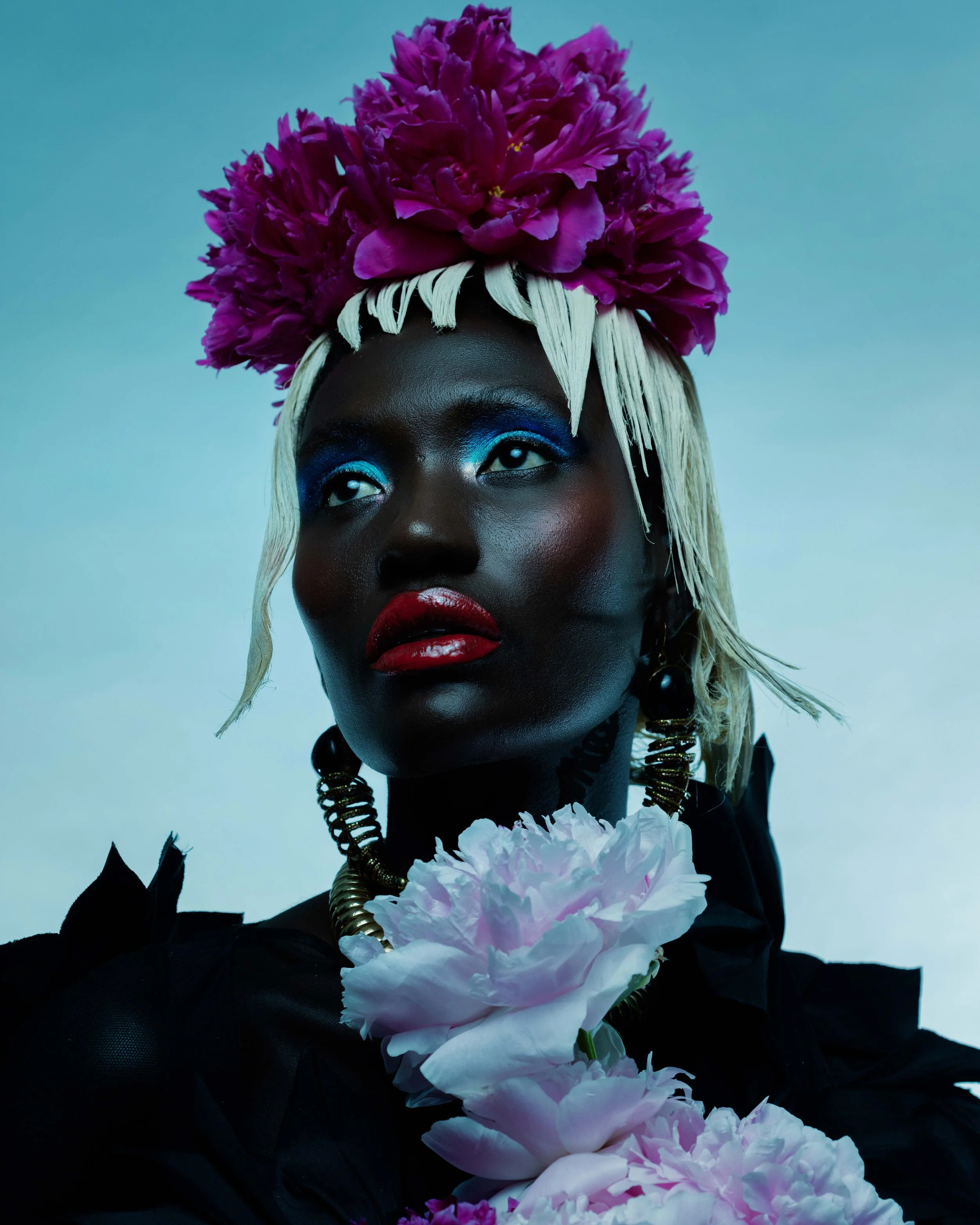 A woman with dark skin and blonde hair, wearing vibrant makeup including blue eyeshadow and red lipstick, adorned with a headpiece of purple flowers and holding pink peonies.