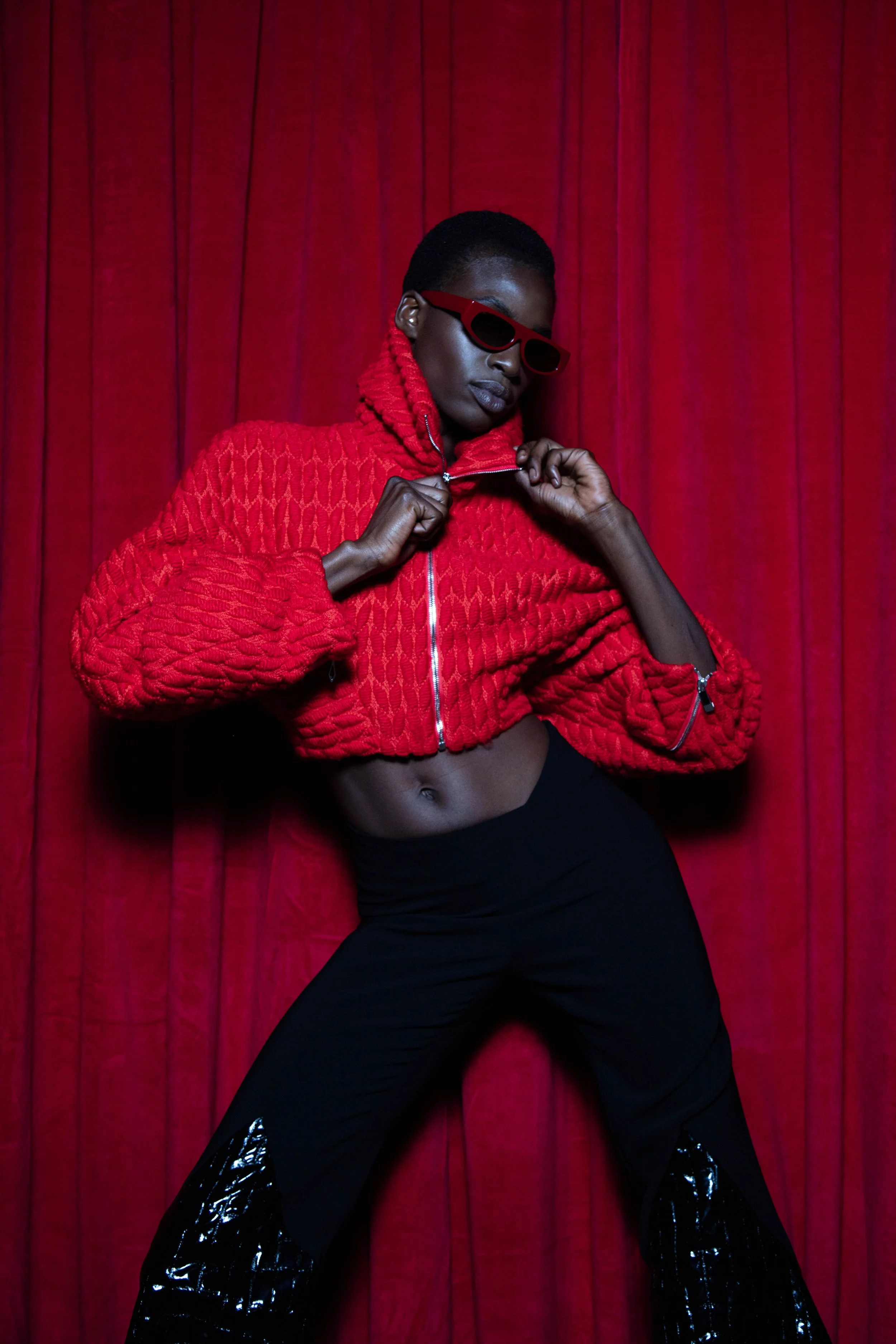 Model wearing a red quilted jacket and black pants with shiny black accents, posing against red velvet curtains.