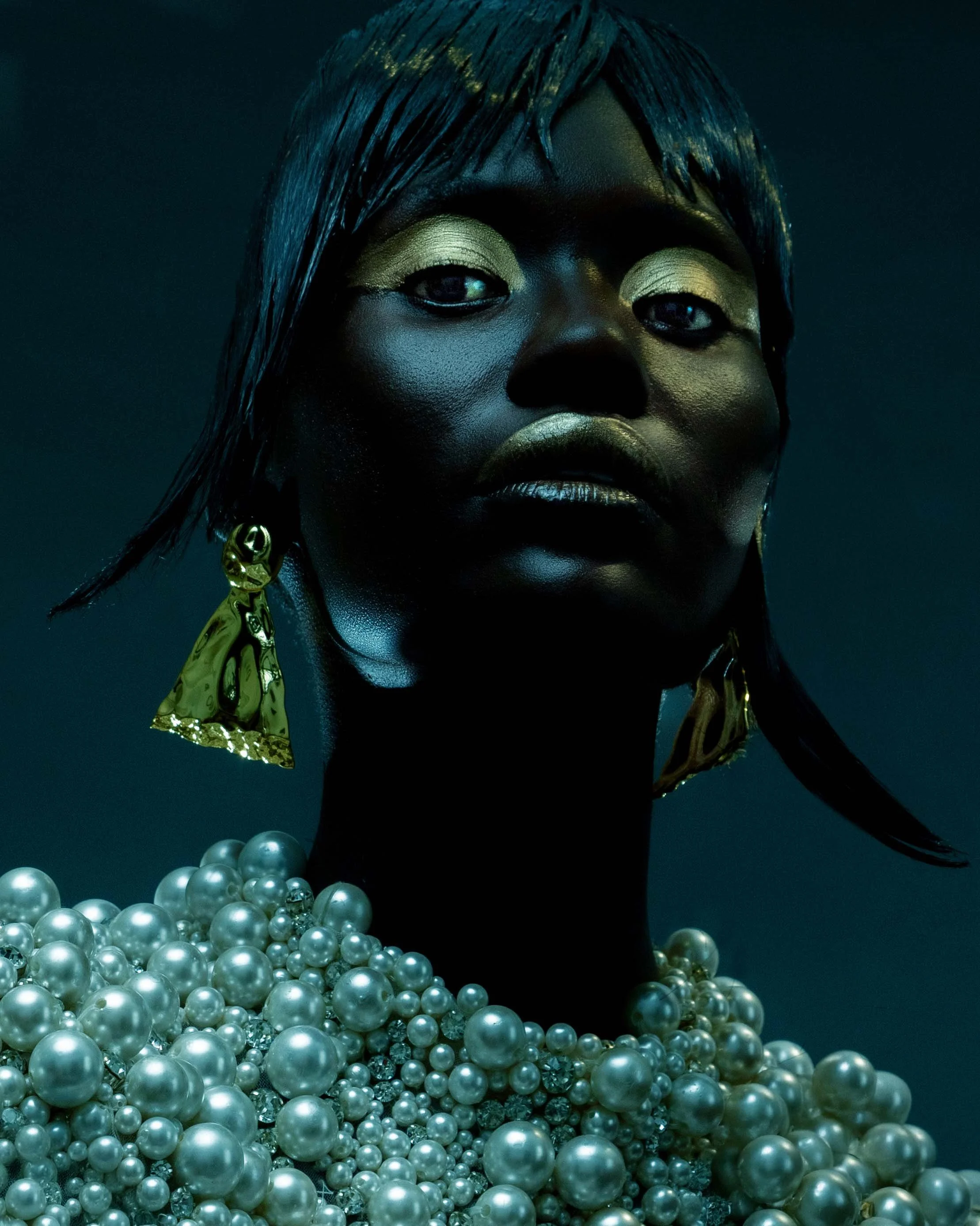 Close-up of a woman with black skin wearing gold makeup, large pearl necklace, and gold earrings against a dark background.