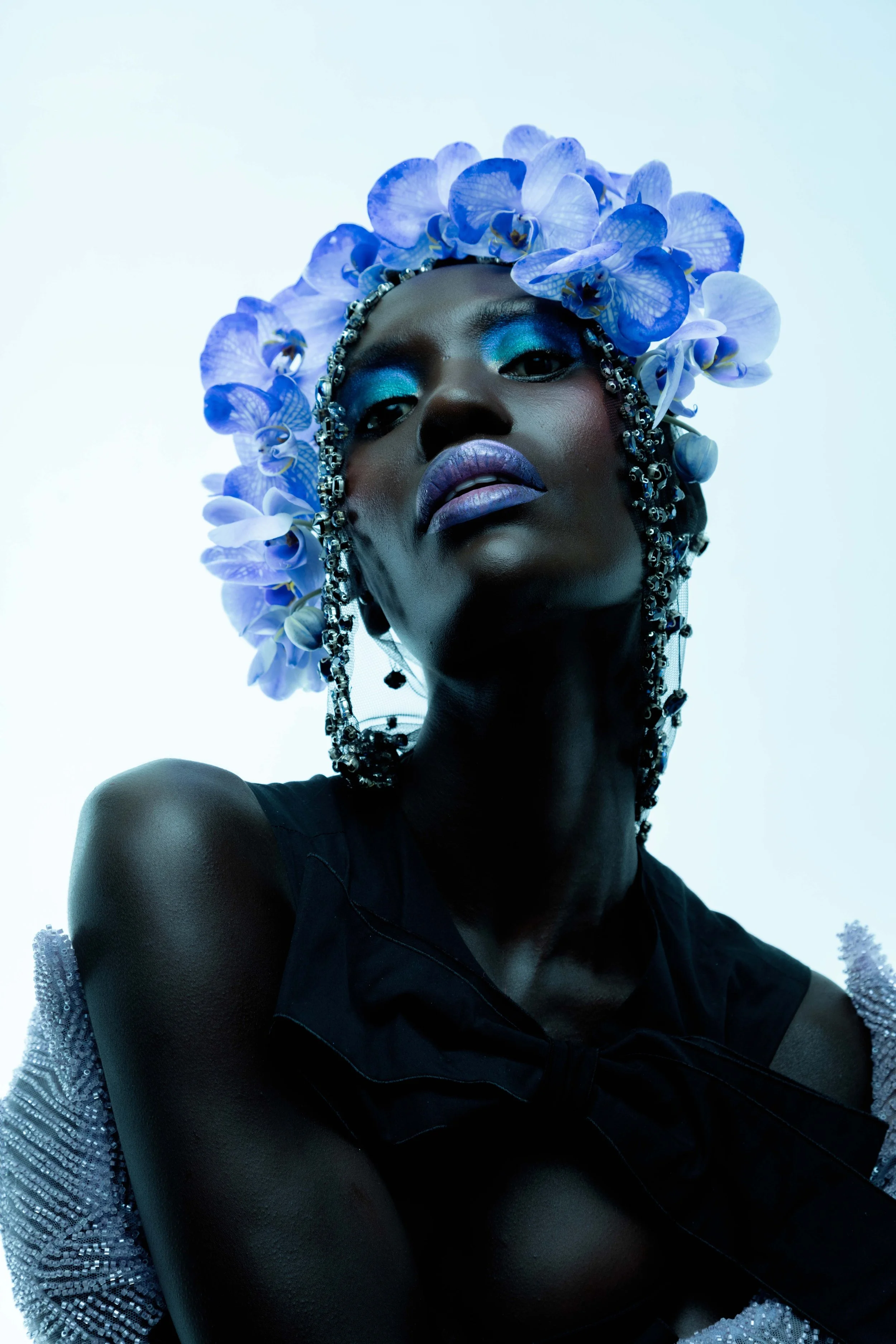 A woman with dark skin wearing a headpiece made of blue orchids, with makeup featuring blue eyeshadow and purple lipstick, against a light background.