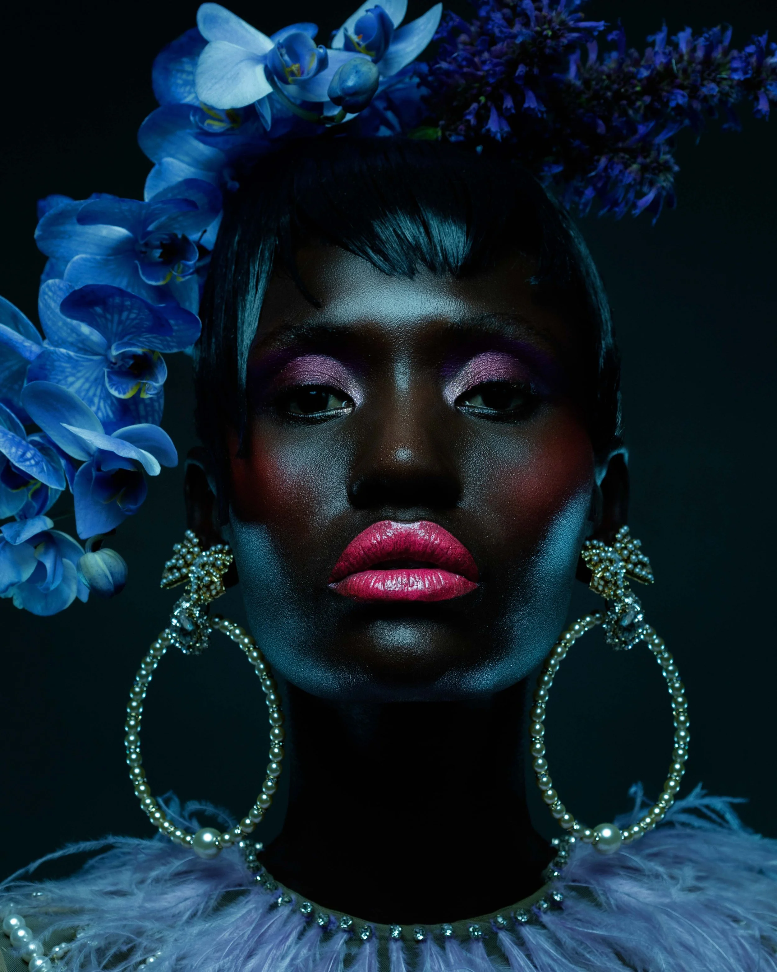 Portrait of a woman with dark skin, bold makeup, and large pearl jewelry, surrounded by blue flowers.
