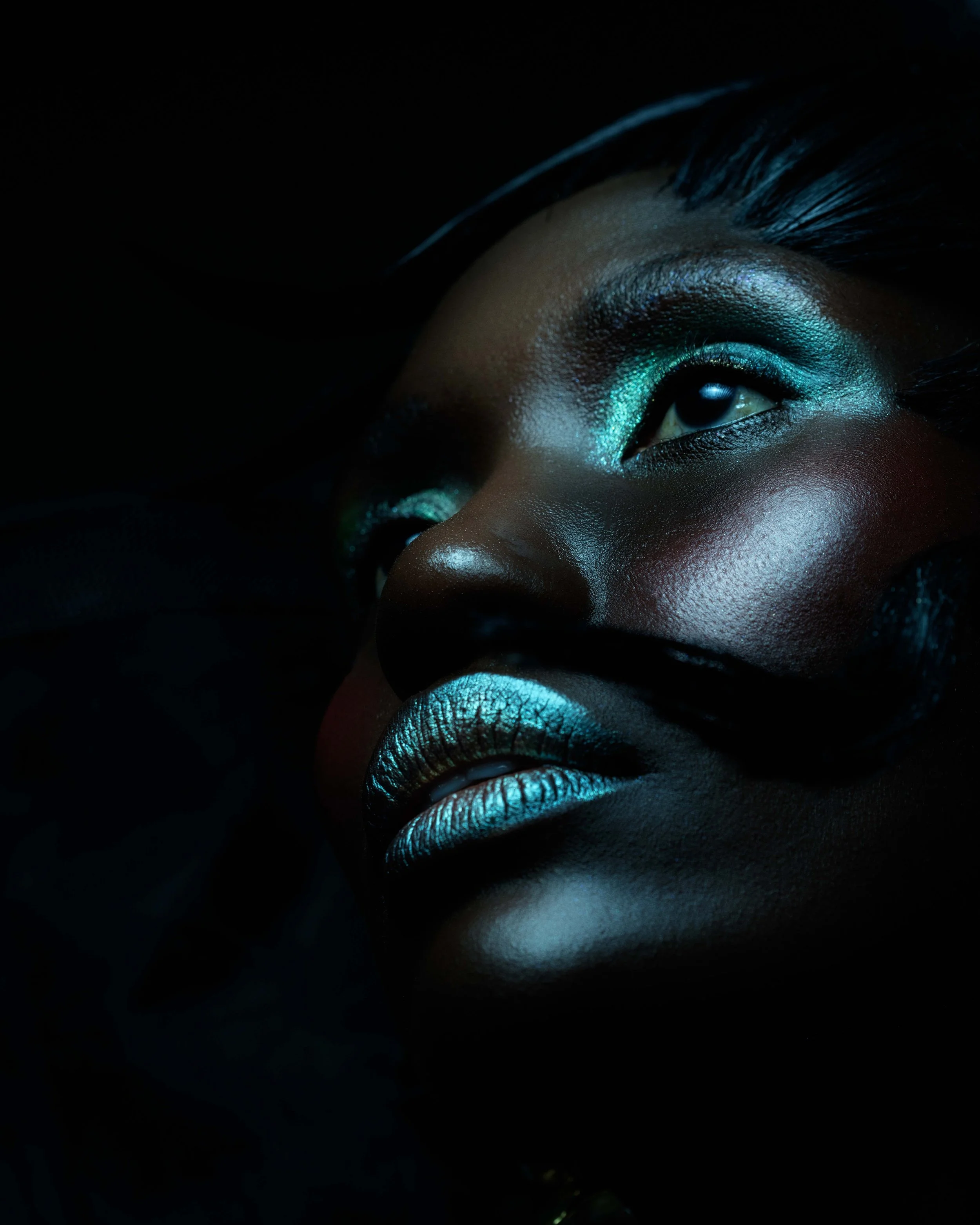 Close-up of a woman's face with makeup in a dark setting, featuring metallic teal eyeshadow and matching lipstick.