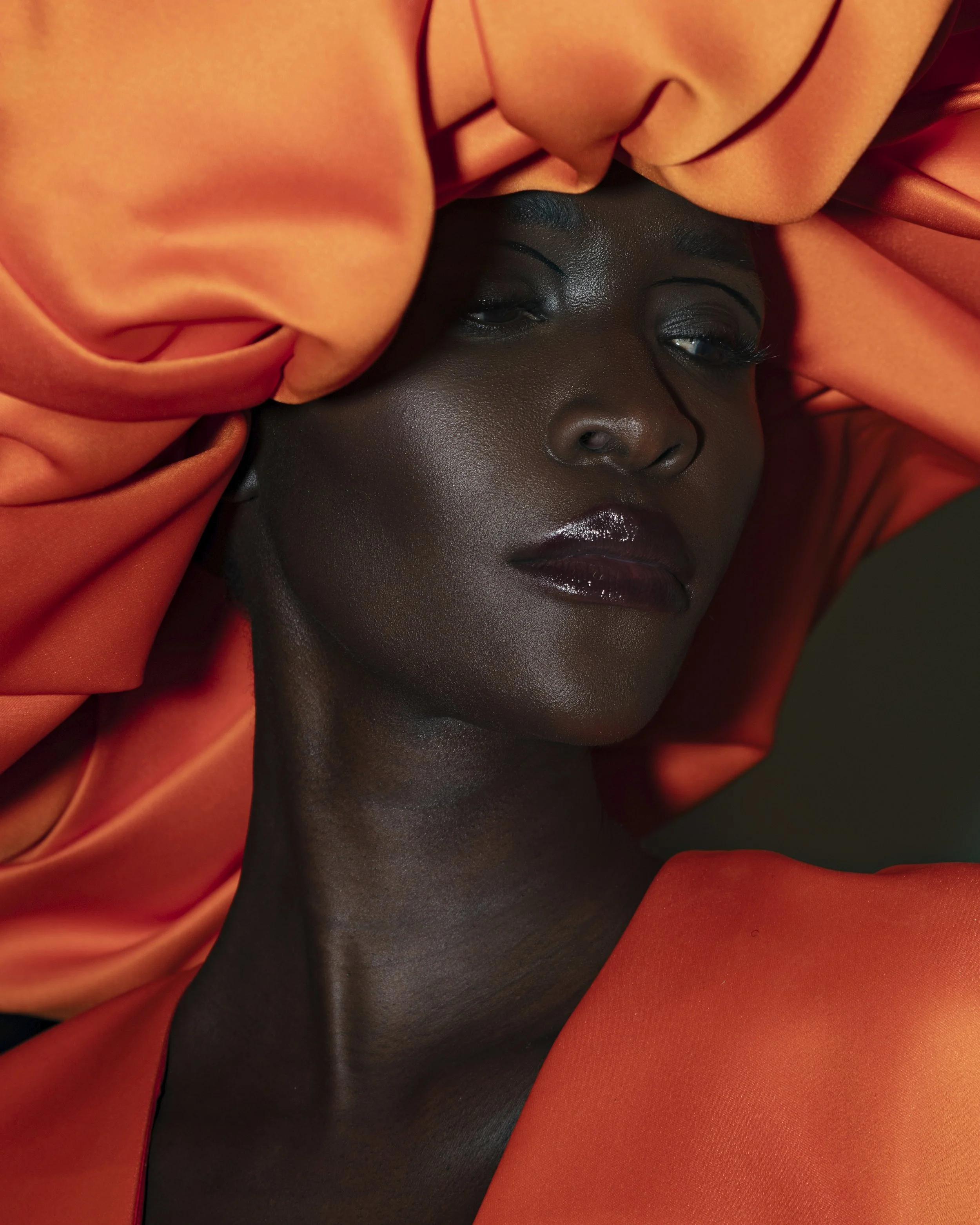 Close-up portrait of a woman with dark skin, short hair, and subtle makeup, framed by flowing orange fabric.