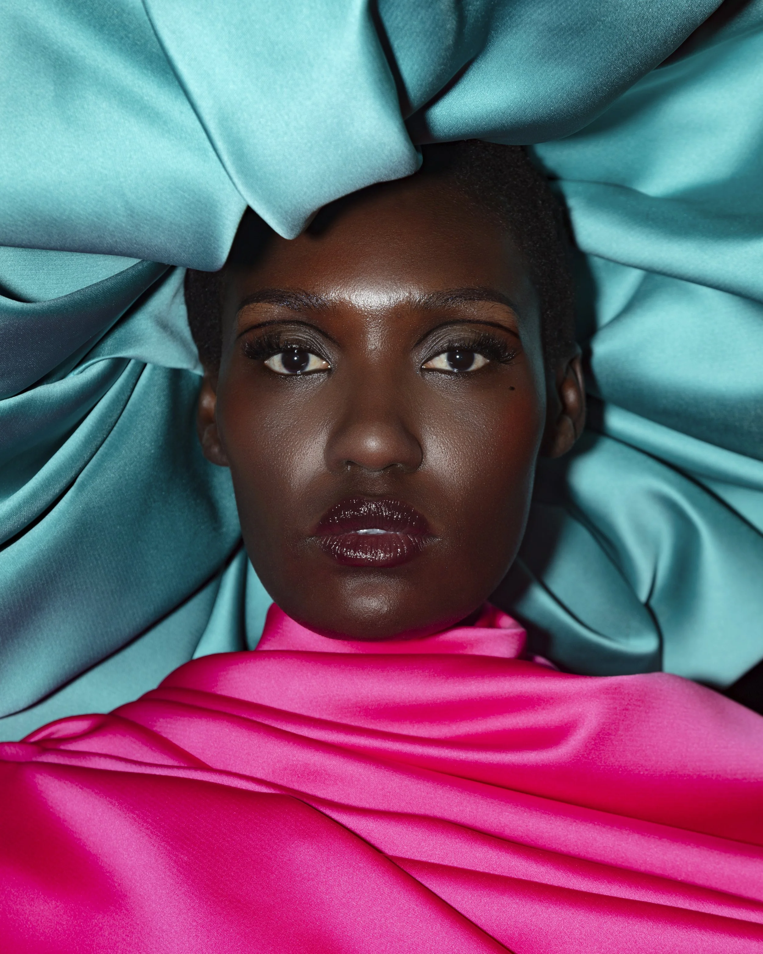 A woman with dark skin and short hair lying on blue satin fabric, wearing bright pink satin garments.
