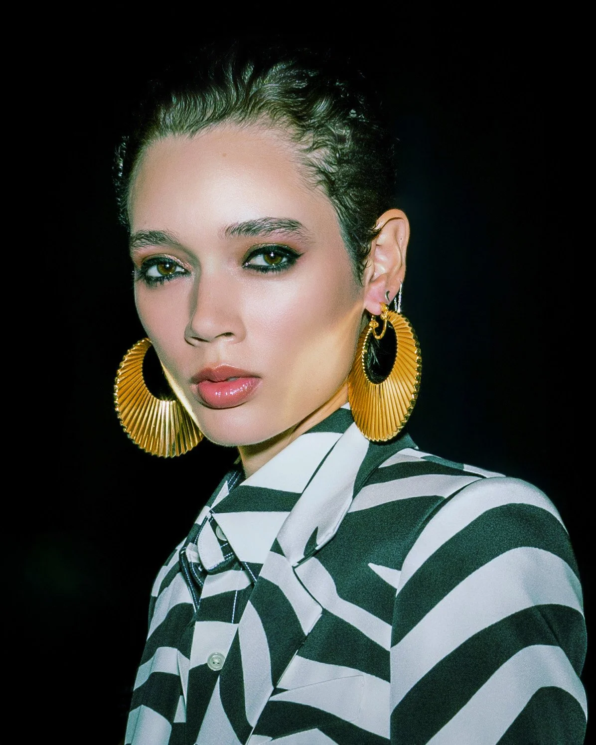 A woman with dark, slicked-back hair, wearing large gold hoop earrings and a black and white striped jacket against a black background.