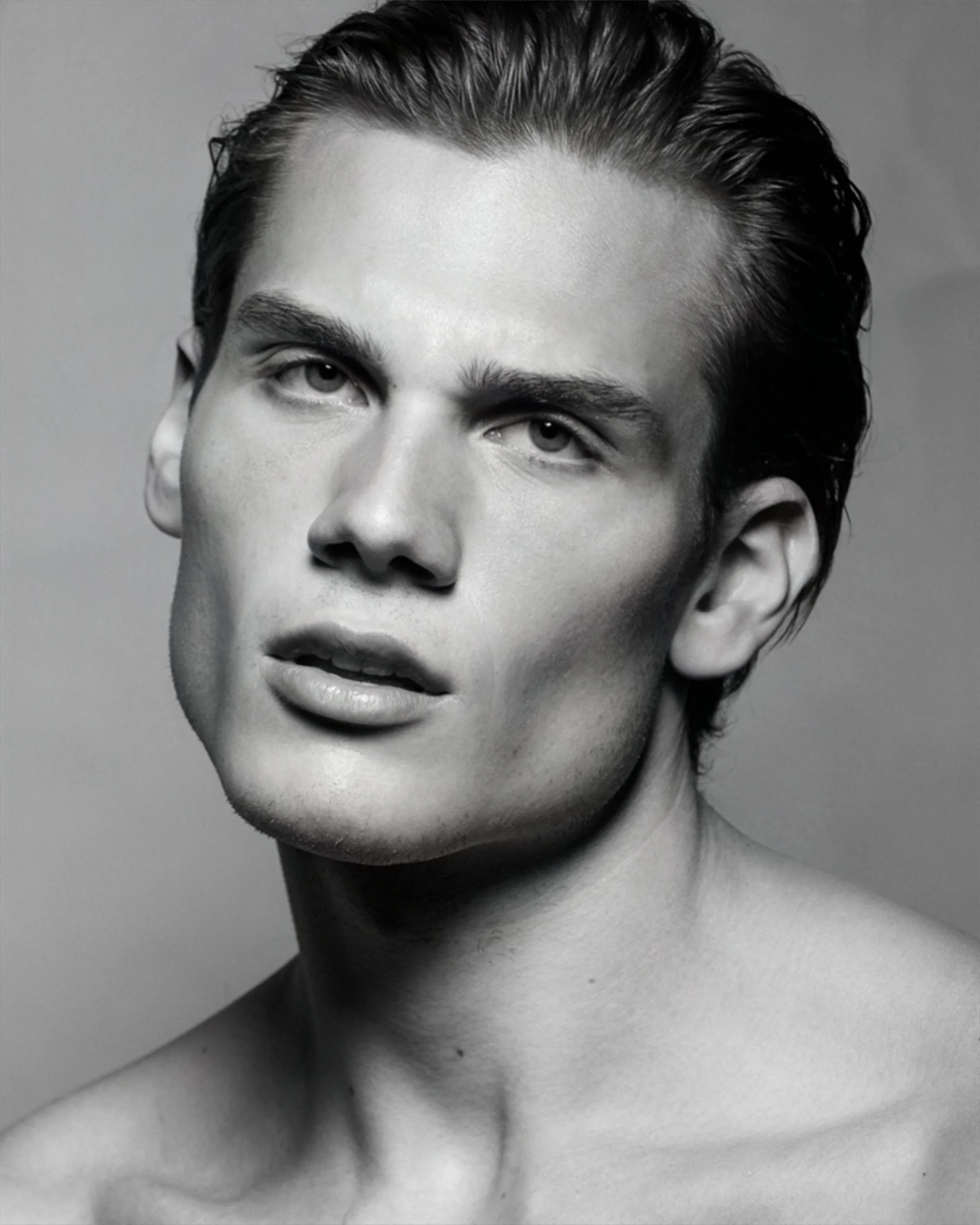 Black and white portrait of a person with short, slicked-back hair, high cheekbones, and full lips, looking slightly to the side with a serious expression.