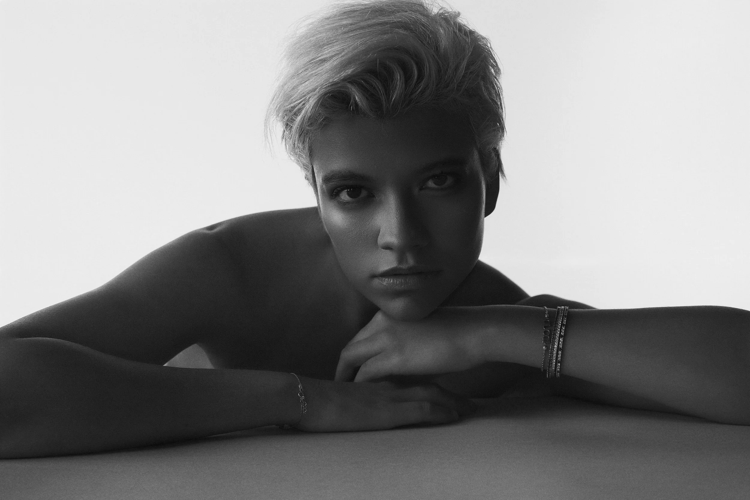A black and white photograph of a woman with short, light-colored hair, resting her head on her crossed arms on a flat surface, looking directly at the camera with an intense expression, wearing bracelets on her wrist.
