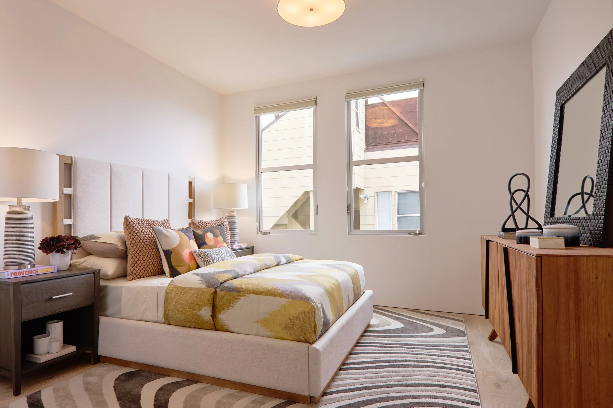 A modern bedroom with a bed, two nightstands with lamps, a dresser with decorative objects, and windows with view outside.