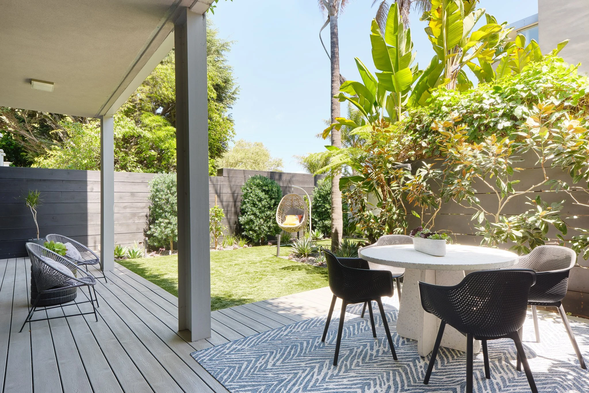 A backyard patio with patio furniture, a round table with black chairs, a hanging chair with a yellow pillow, green plants, and a wooden privacy fence under a bright sunny sky.