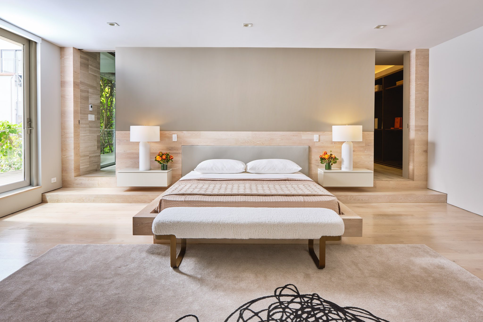 Modern bedroom with a bed, two nightstands with lamps and flower arrangements, large window with sliding glass door showing greenery outside, and a beige area rug.