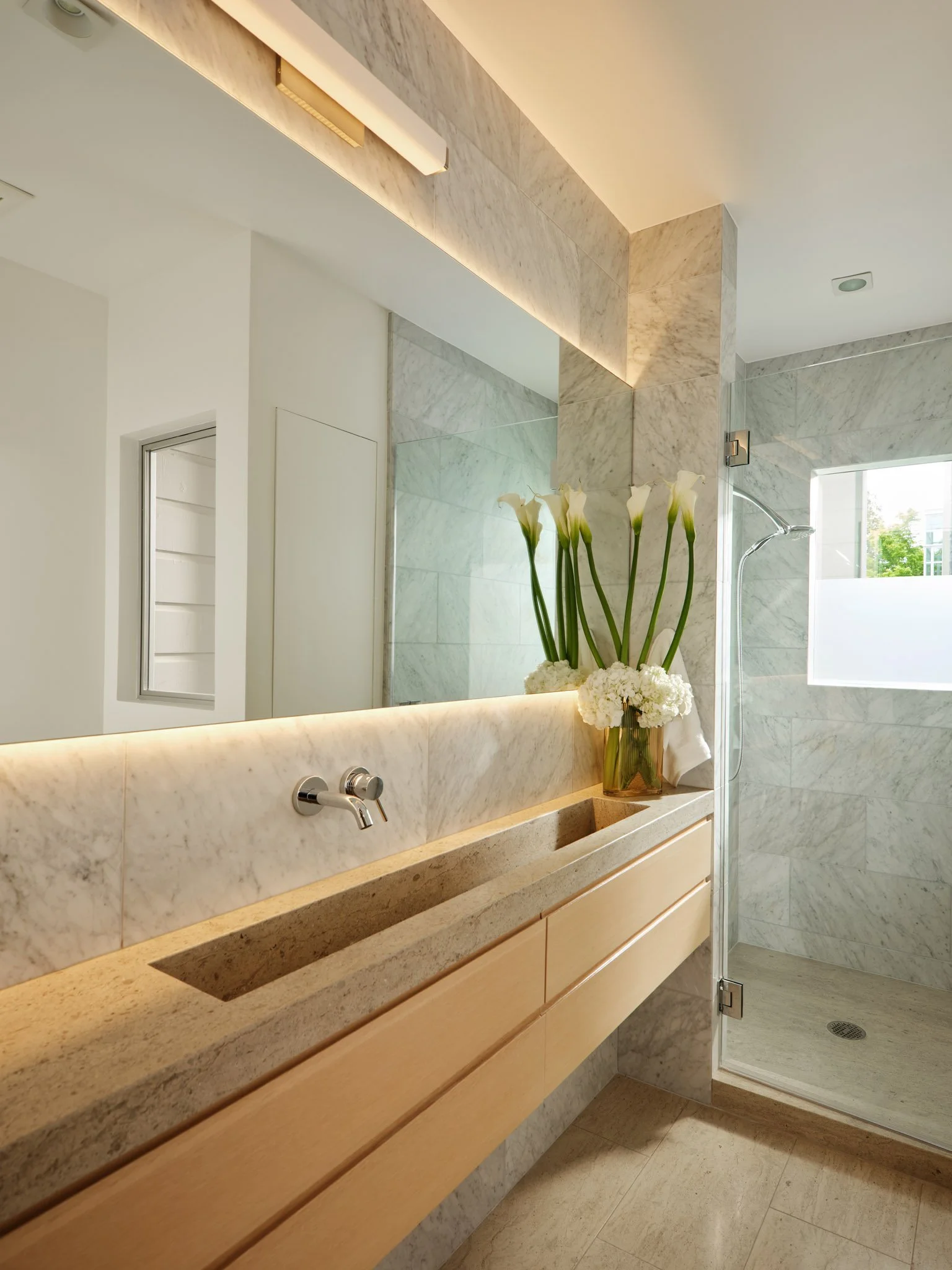 Modern bathroom with beige marble countertops, a large mirror, a vase with white calla lilies and hydrangeas, and a walk-in shower with marble walls and a glass door.