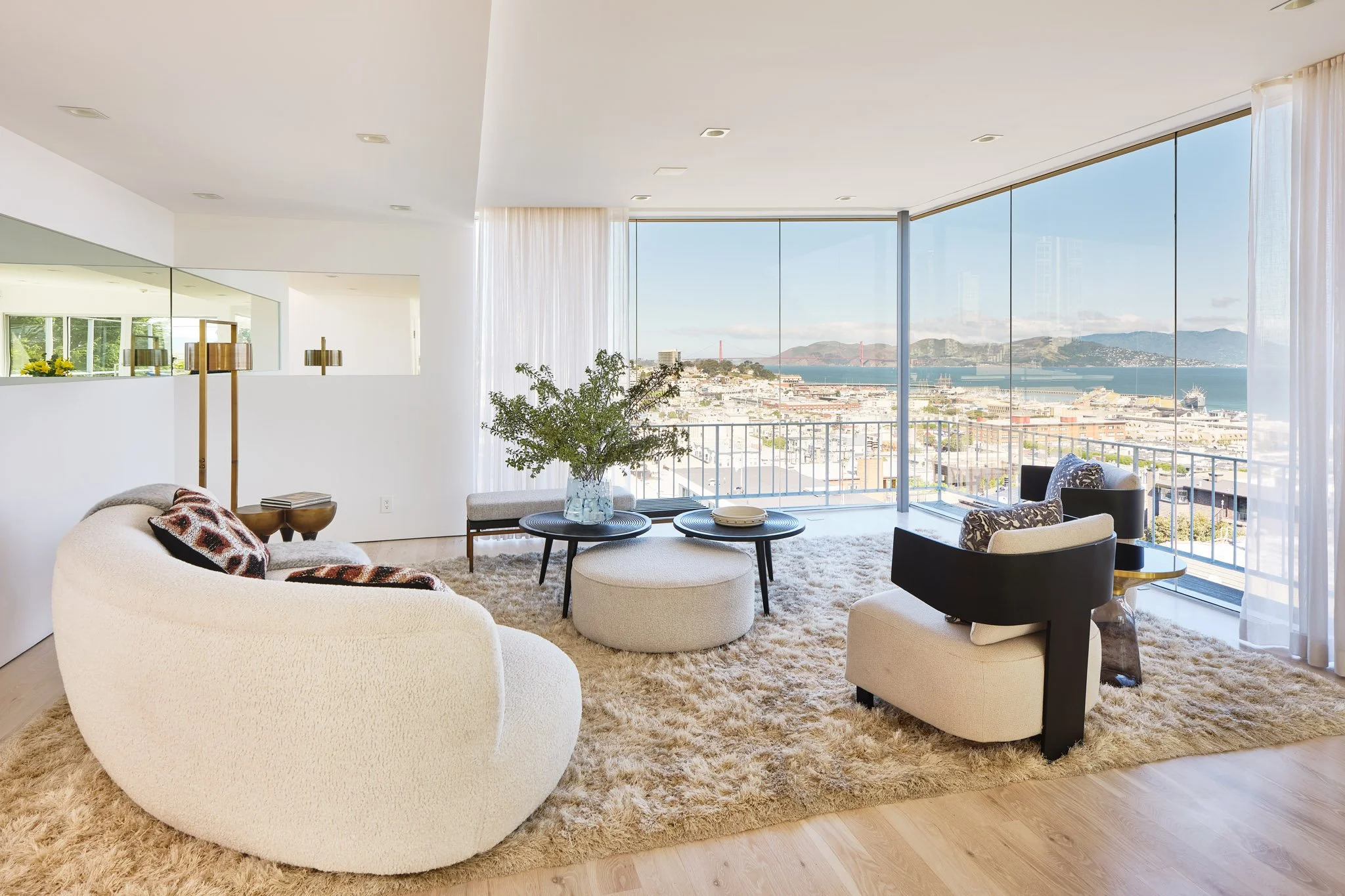 Living room with large glass windows overlooking a city and water, beige carpet, white curved sofa, black and beige armchairs, round coffee tables, decorative pillows, and an indoor plant in a glass vase.