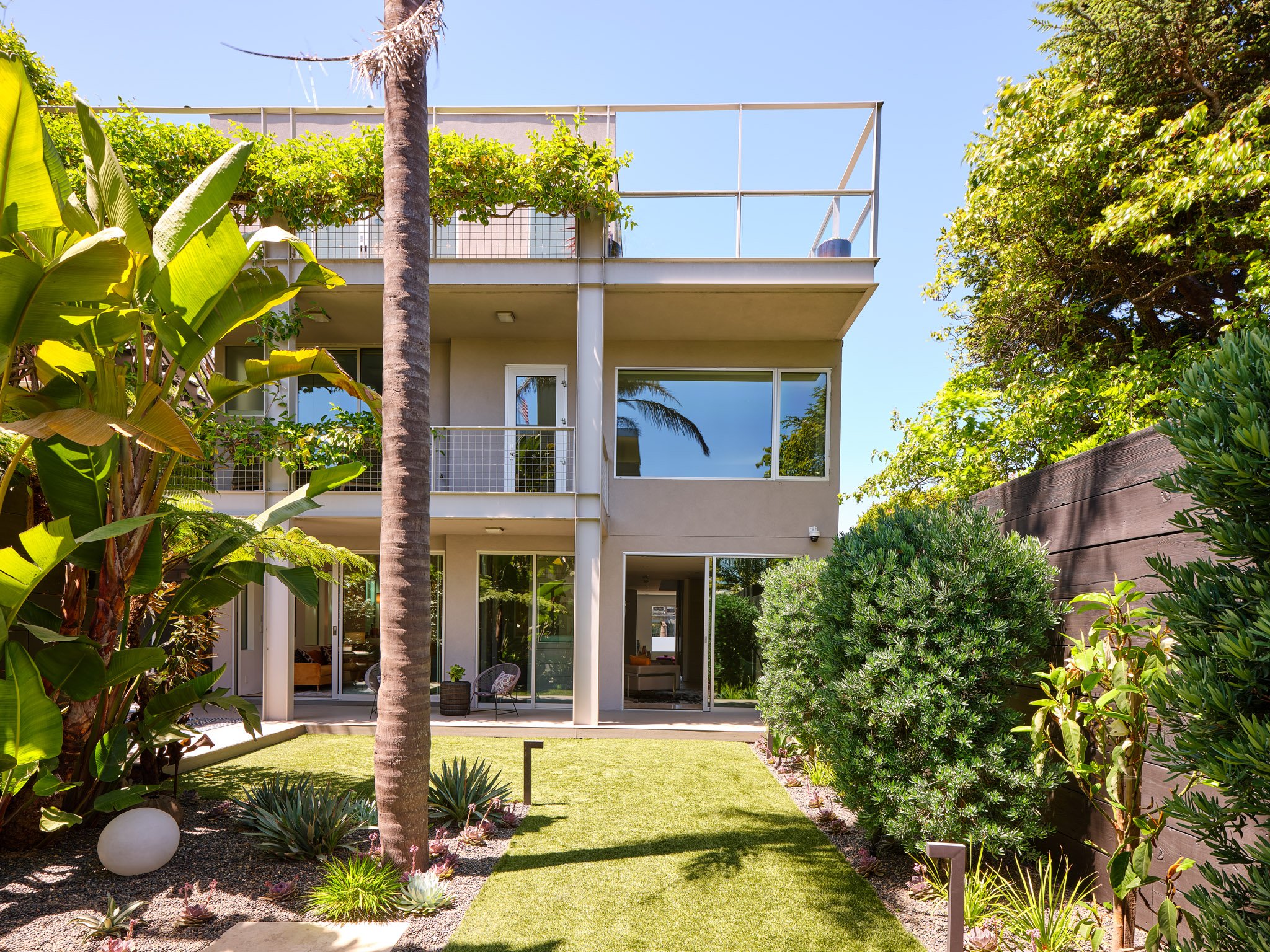 View of a modern three-story house with large glass sliding doors and balconies, surrounded by lush tropical plants and a well-maintained green lawn, on a sunny day.