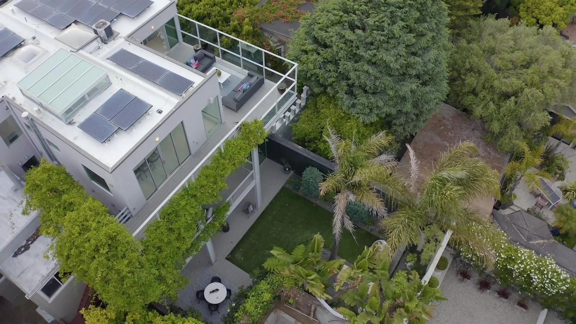 Aerial view of a modern multi-story house with a rooftop deck, solar panels, outdoor seating, and surrounding lush green trees and plants.