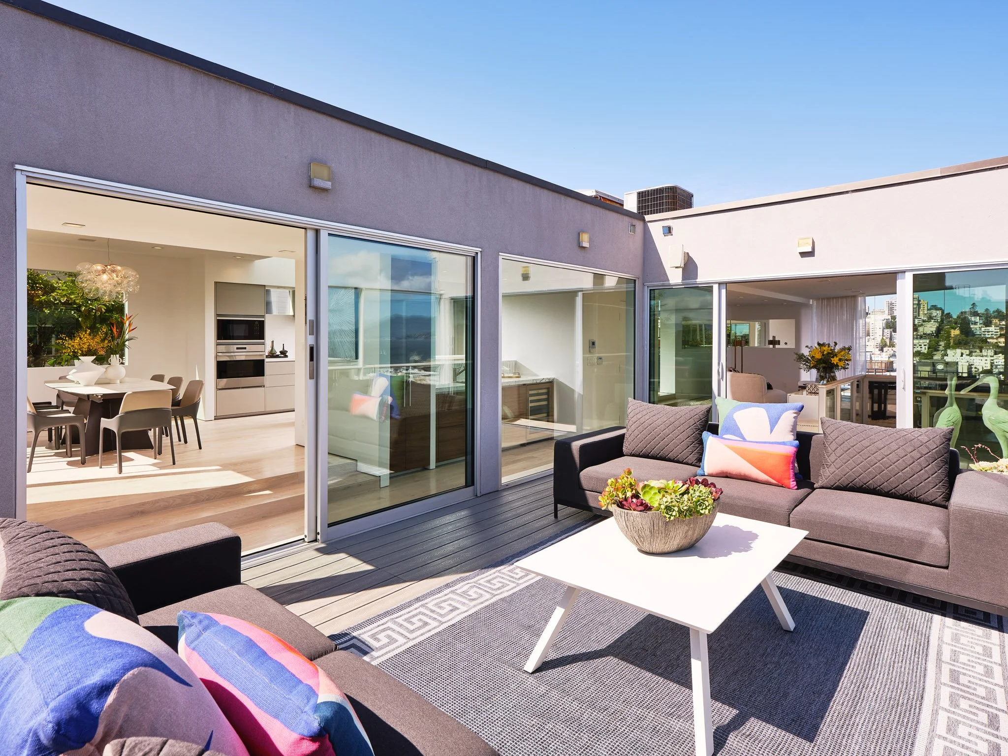 Outdoor patio area with lounge chairs, cushions, a white coffee table with a flower arrangement, and glass sliding doors leading inside a modern apartment with dining and kitchen areas visible.