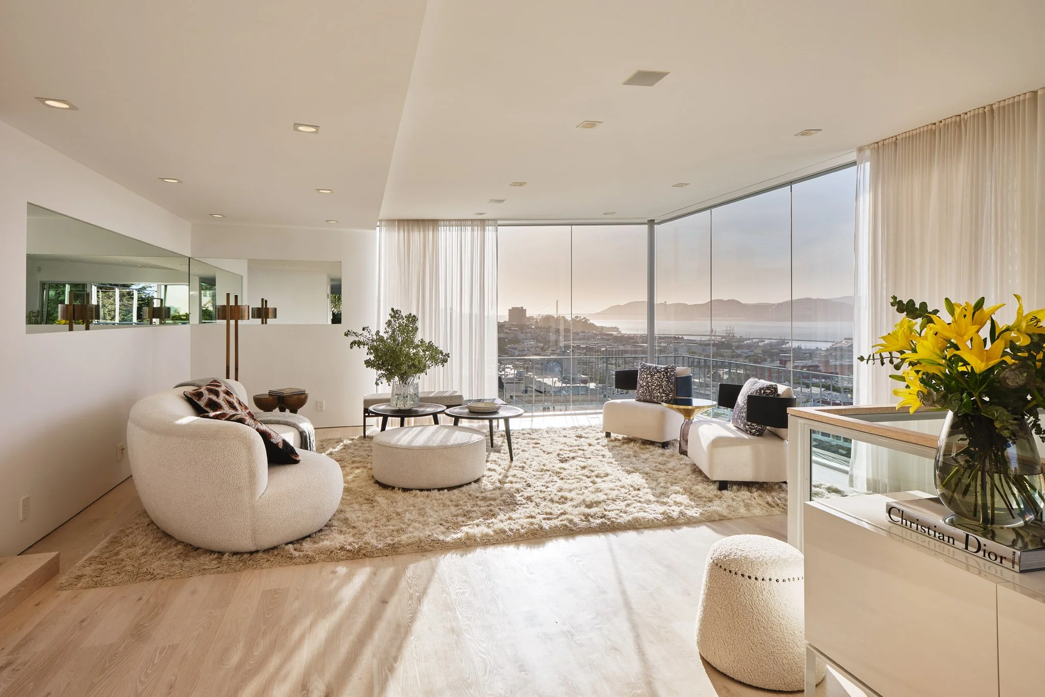 Modern living room with large floor-to-ceiling windows overlooking a cityscape, featuring white curved sofas, a fluffy beige rug, coffee tables, and decorative flowers.