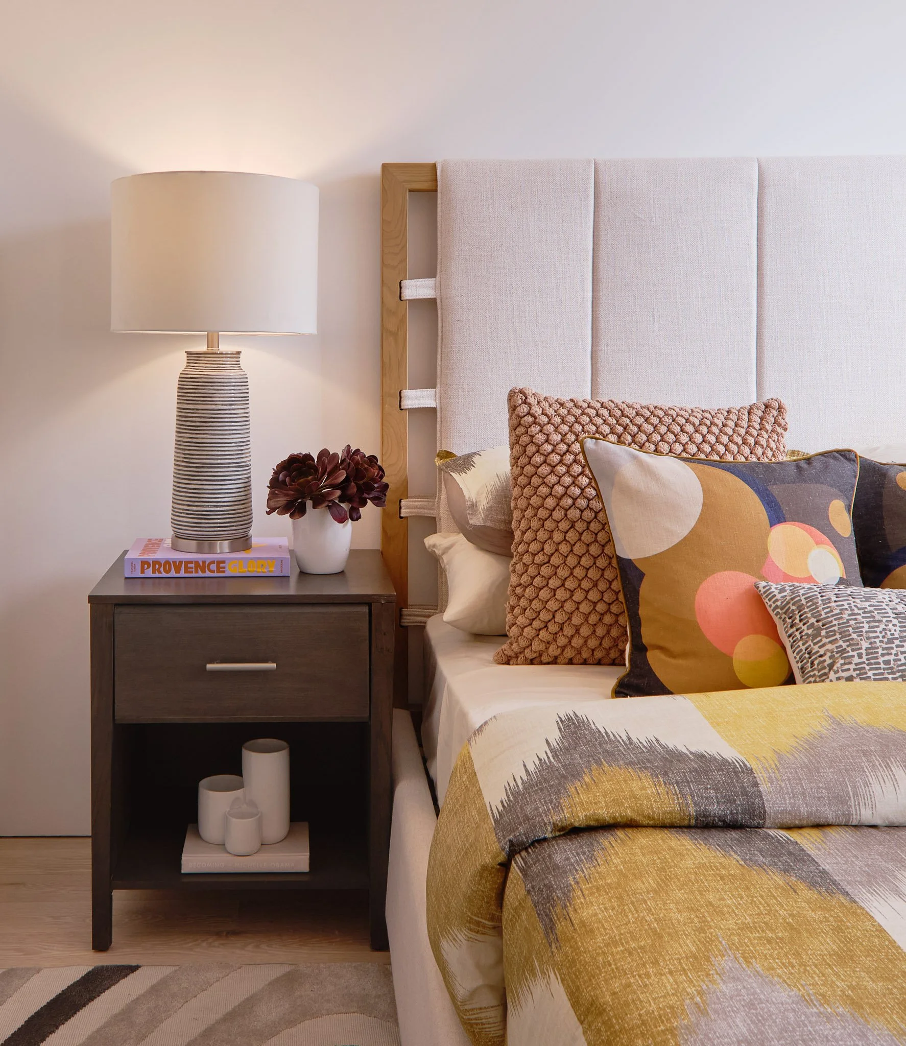 Side of a bedroom with a nightstand, table lamp, flower, and colorful pillows on the bed.