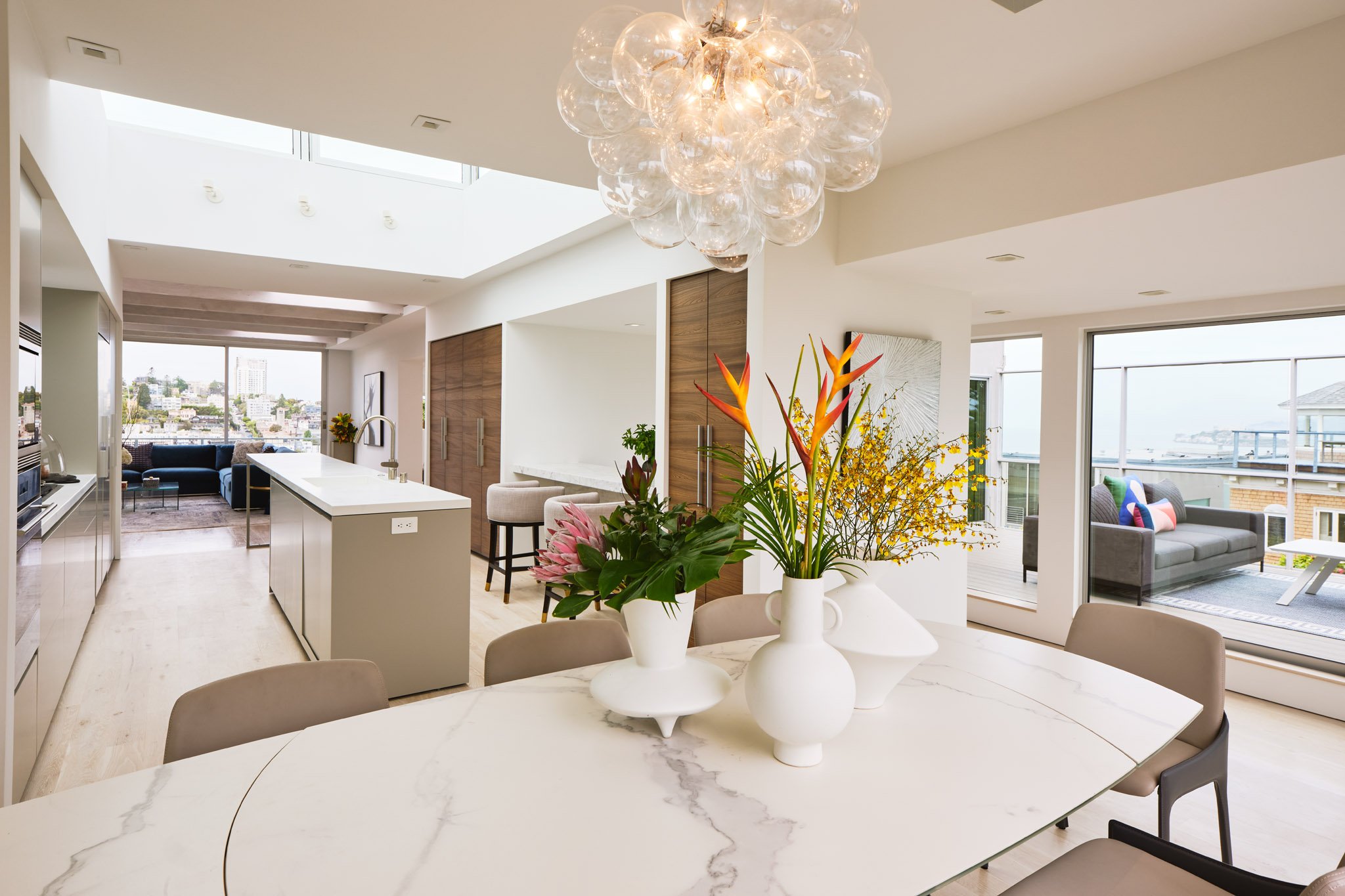 Modern open-concept living space with a marble dining table with vases and flowers, a kitchen island, and living area with large windows and outdoor balconies.