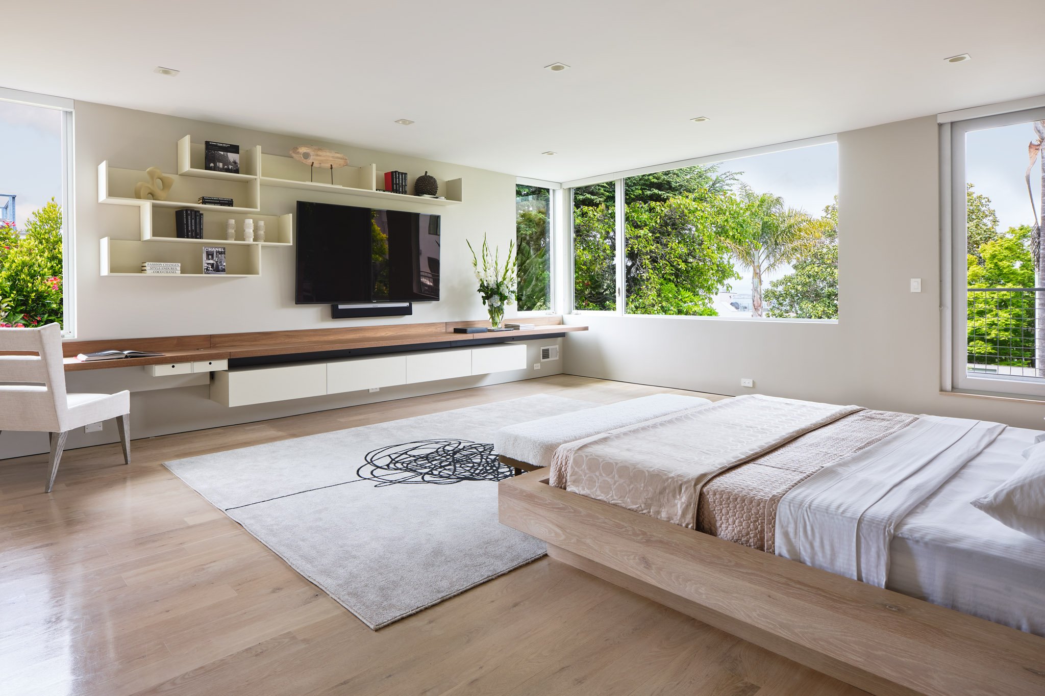 Modern bedroom with large windows showing greenery, a low wooden bed with white bedding, a light-colored rug with abstract black lines, a built-in white entertainment unit with a wall-mounted TV, a vase of flowers, a white chair, and minimalist float
