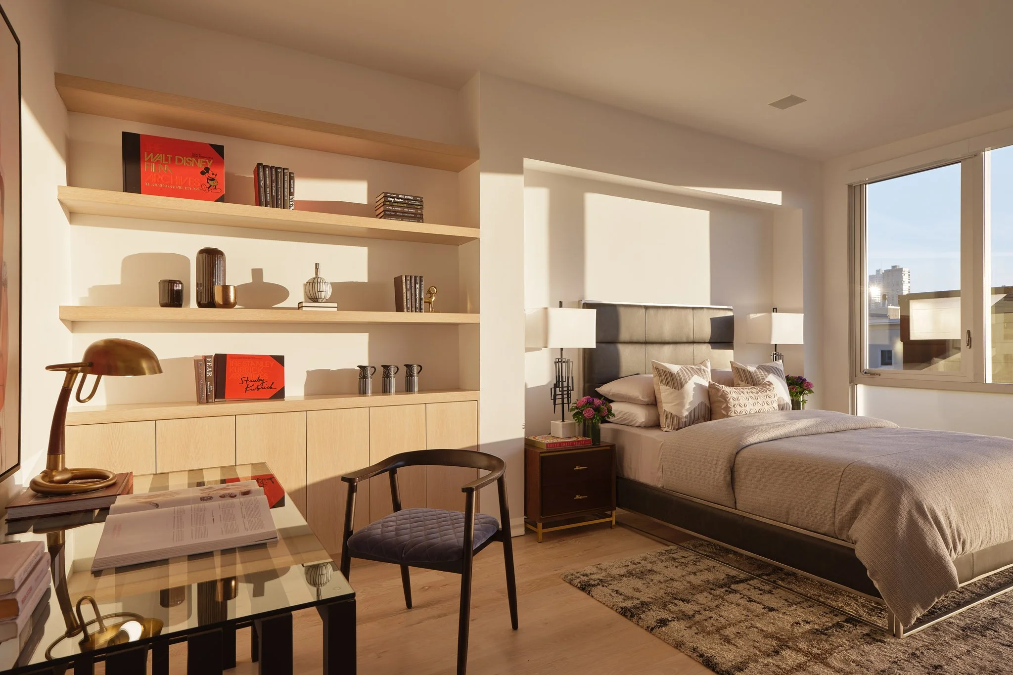 Bright bedroom with a large bed, decorated with pillows, next to a window showing a cityscape, with nightstands, lamps, and a textured rug on the wooden floor. An open shelf with books and decor items is on the left.