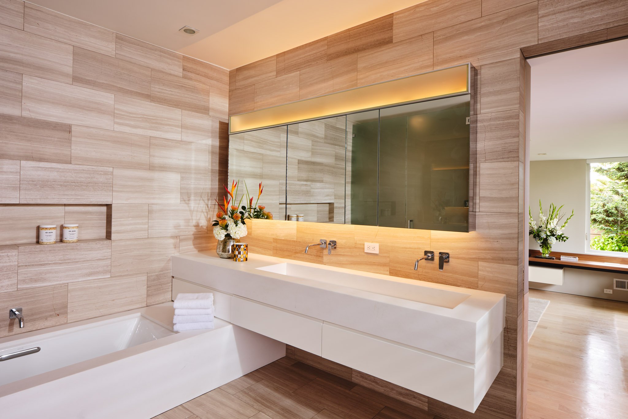 Modern bathroom with wooden wall tiles, a large mirror, a white countertop with a built-in sink, a bathtub with towels, and a vase of flowers, with natural light coming in from a window