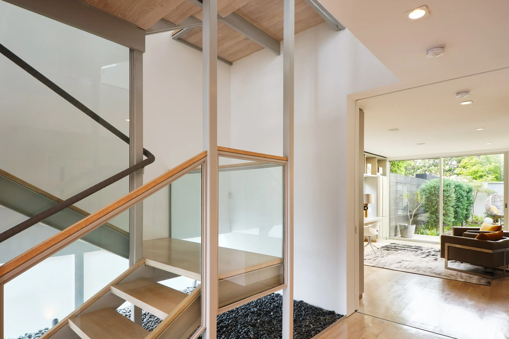 Modern interior featuring a staircase with glass panels and wooden trim, next to a white wall, leading to a bright living room with large windows, greenery visible outside, and minimalistic furniture.