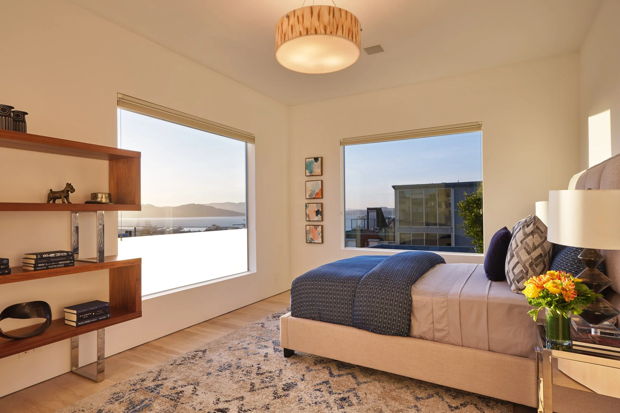 Modern bedroom with large windows showing a view of water and mountains, bed with dark blue bedding, and wooden shelf with decorative items.