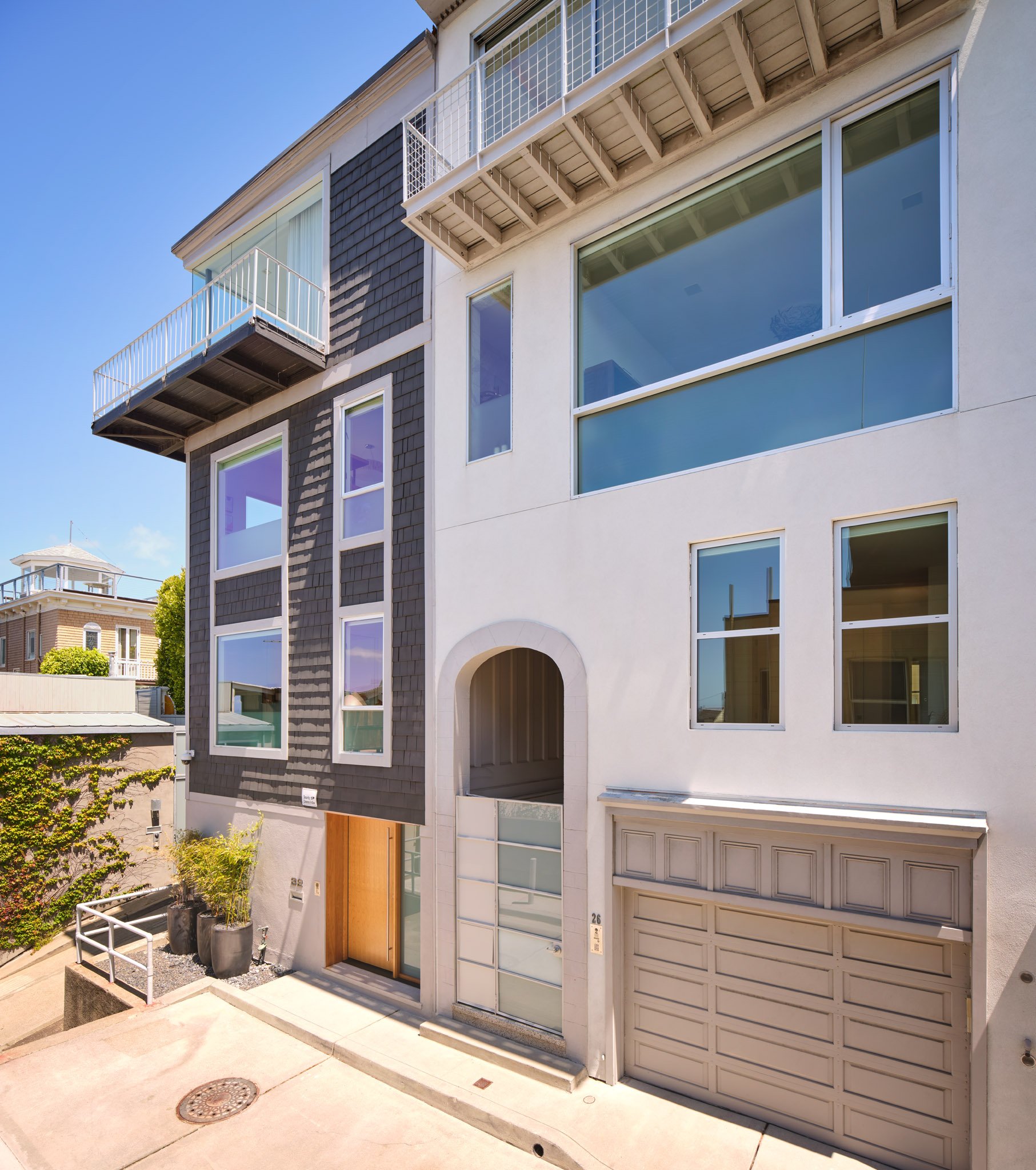 Modern multi-story residential building with large windows, a balcony, and a garage door, situated in an urban area under a clear blue sky.