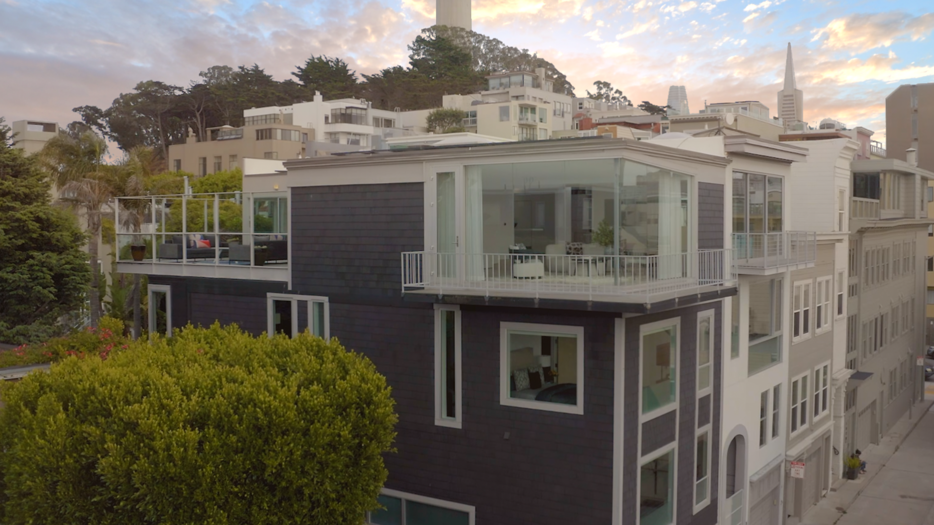 Modern multi-story apartment building with large windows and balconies, set against a backdrop of a hilly cityscape at sunset.