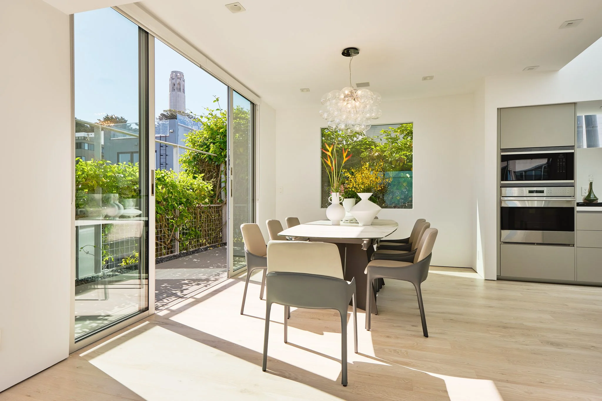 Modern dining area with a round white table, eight beige and gray chairs, decorative vases, and large windows letting in natural light, with outdoor greenery visible.