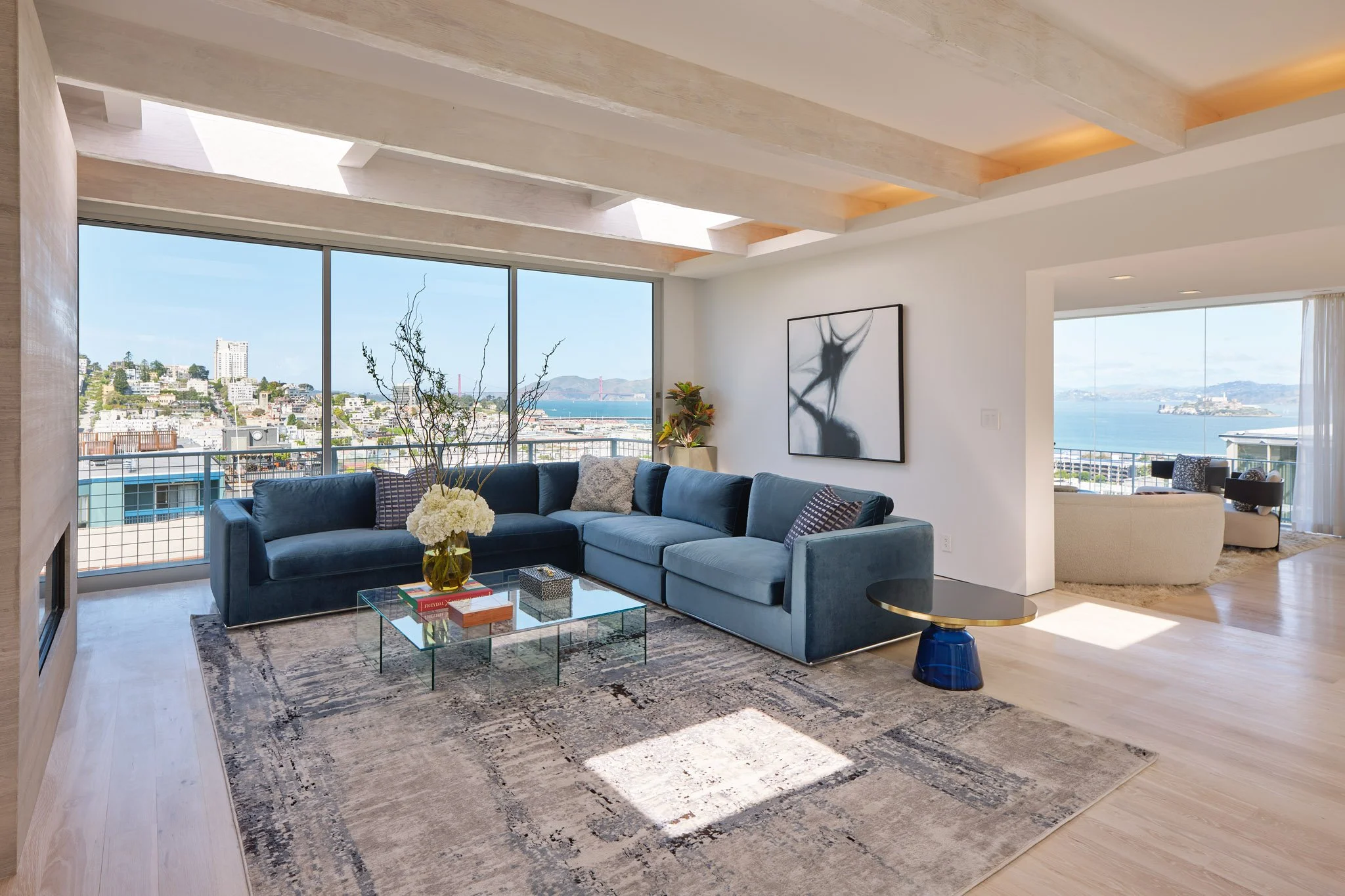 Modern living room with large windows overlooking city and water, featuring a blue sectional sofa, glass coffee table with books and flowers, abstract artwork on white wall, and a cozy white seating area with view.