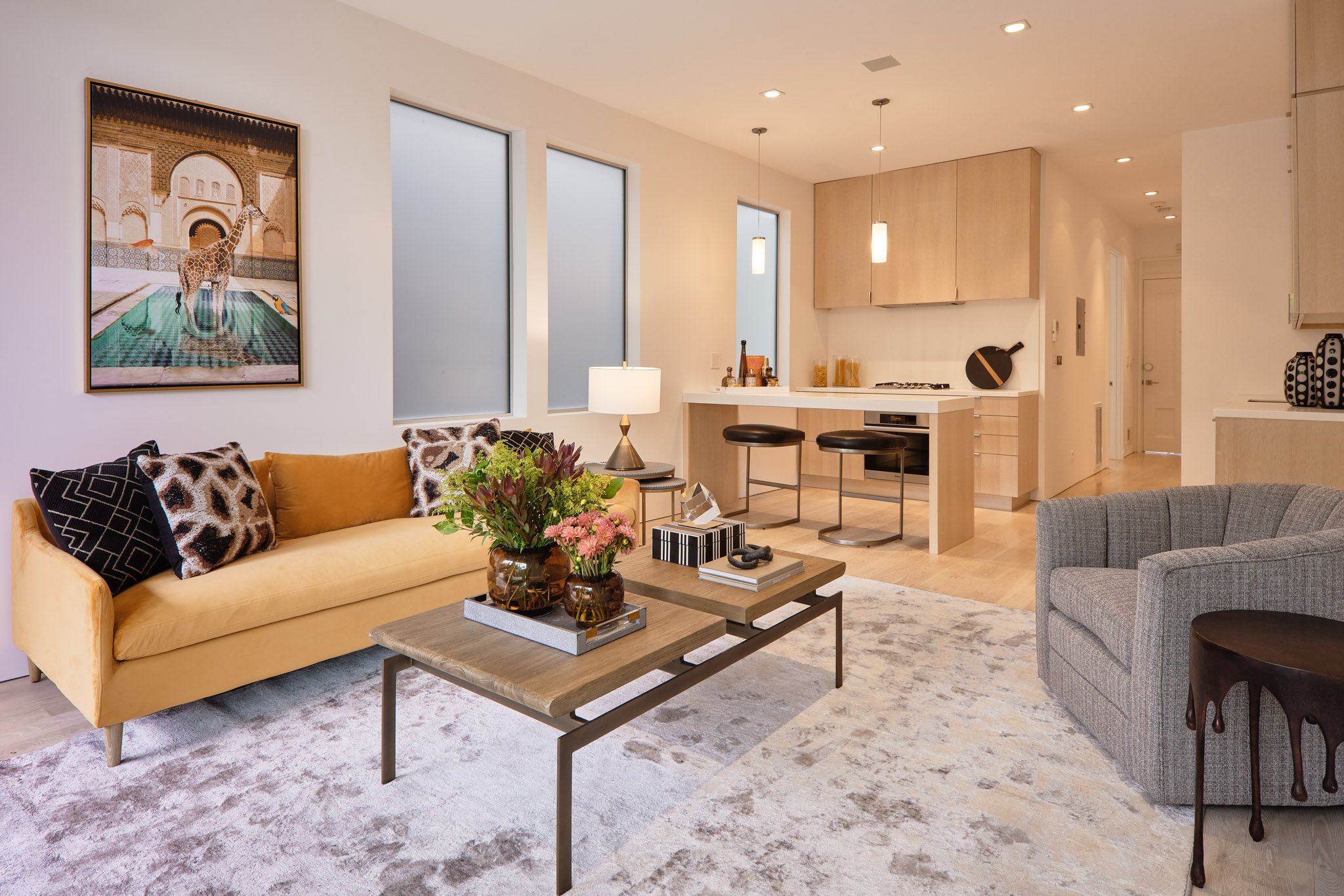 Living room with yellow sofa, patterned pillows, grey armchair, coffee tables with flowers and books, and an open kitchen with light wood cabinets and black bar stools.