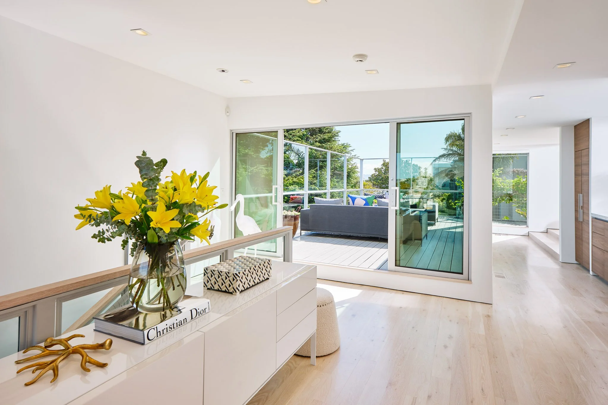 Bright living room with white walls and light wooden floors, leading to an outdoor balcony with seating, glass sliding doors, and greenery outside, decorated with a yellow flower arrangement and modern decor.