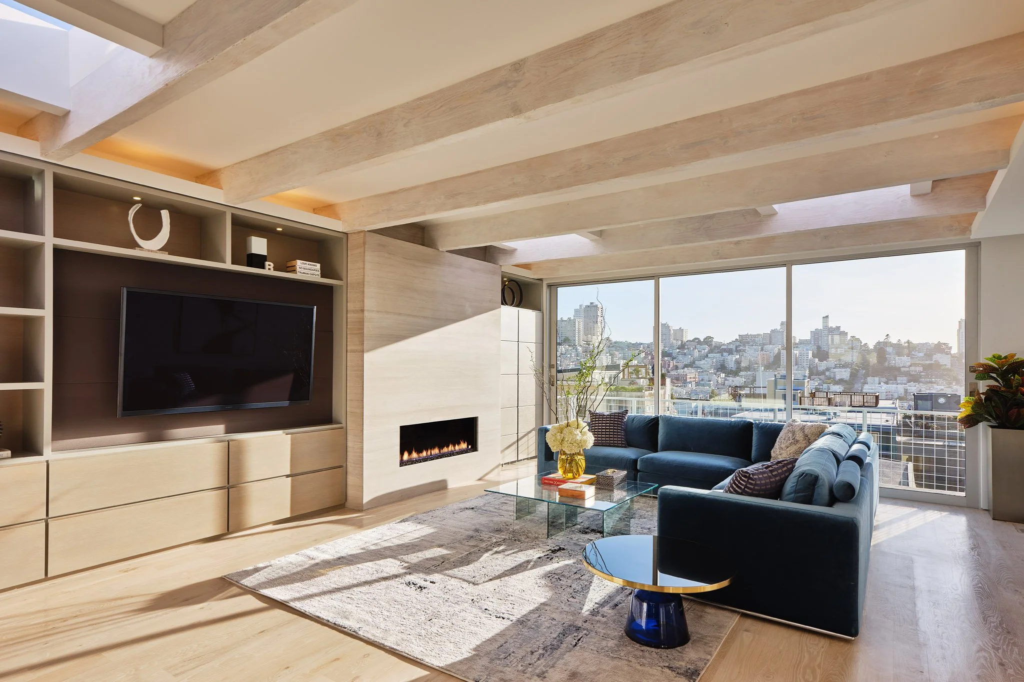 Modern living room with large windows showcasing city view, navy blue sofa, glass coffee table with books and flowers, fireplace, wooden floor, and contemporary decor.