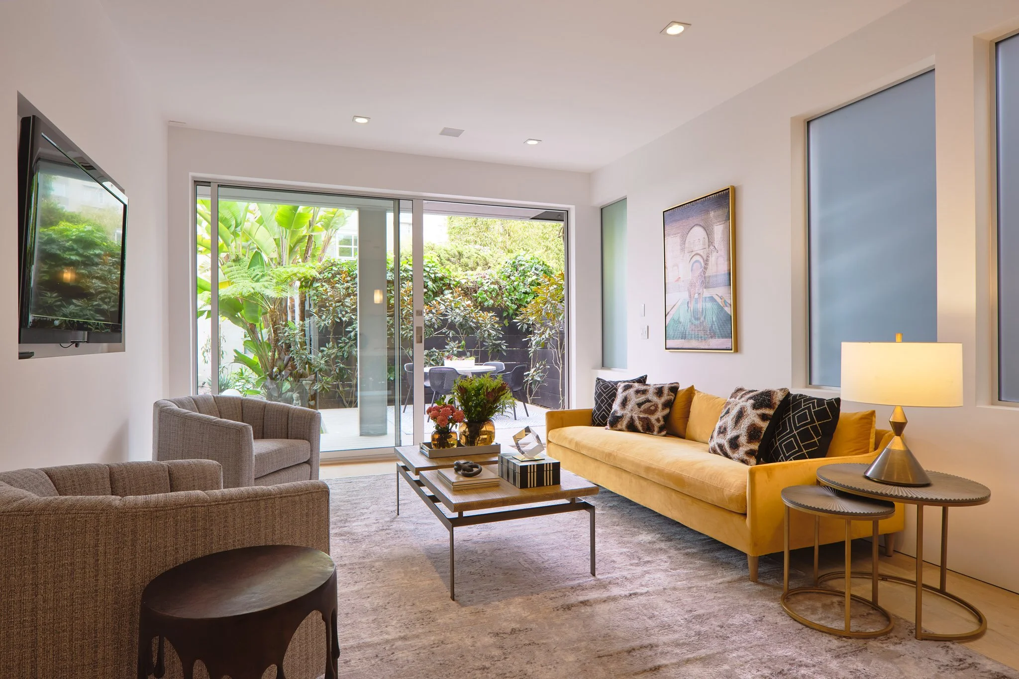 Living room with a yellow sofa, beige armchairs, coffee table, artwork, table lamp, TV, and sliding glass door leading to a patio with greenery.