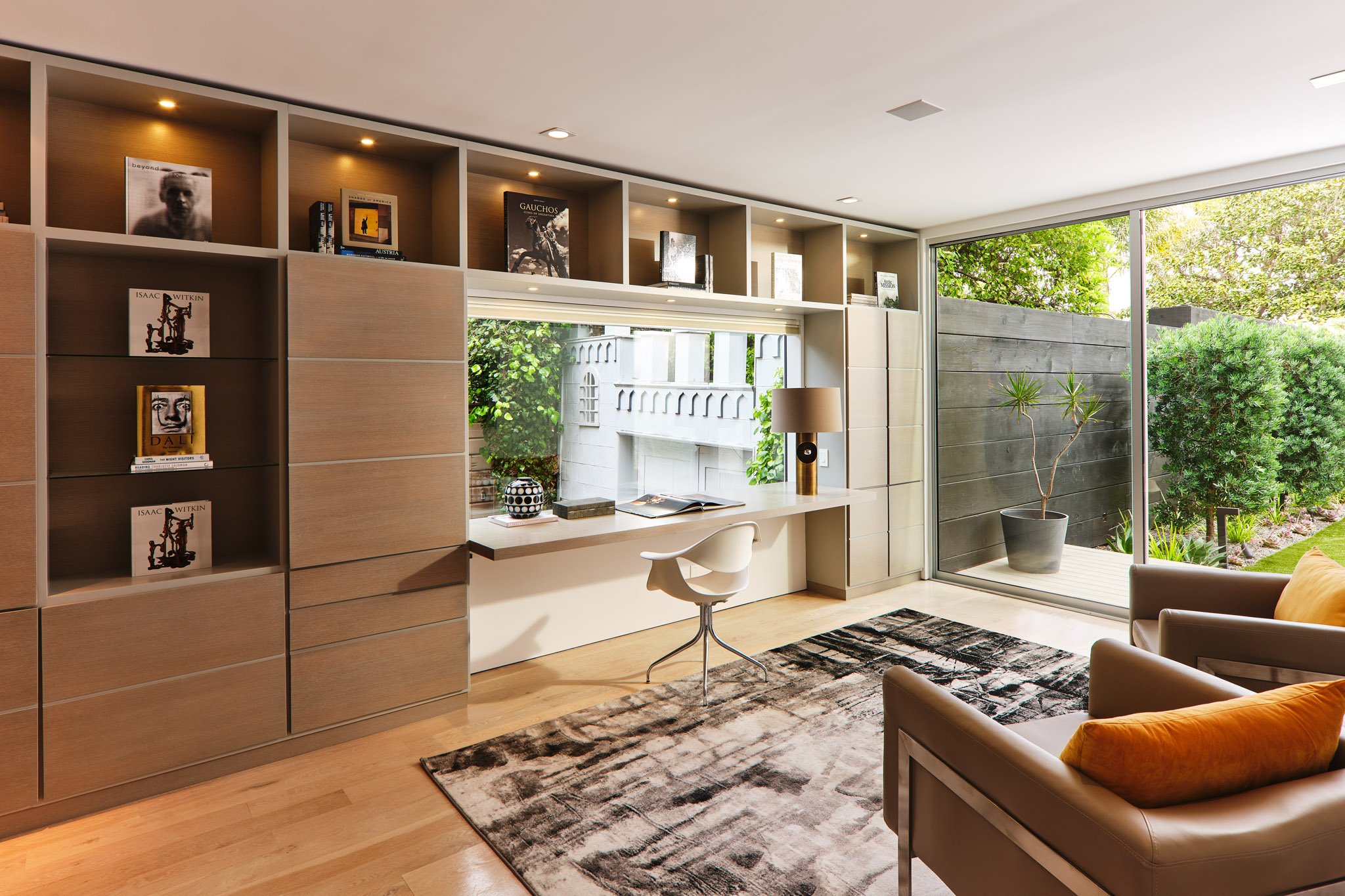Modern living room with built-in bookshelf, desk area, leather armchair with yellow pillow, large window with view of garden, and potted plant outside.