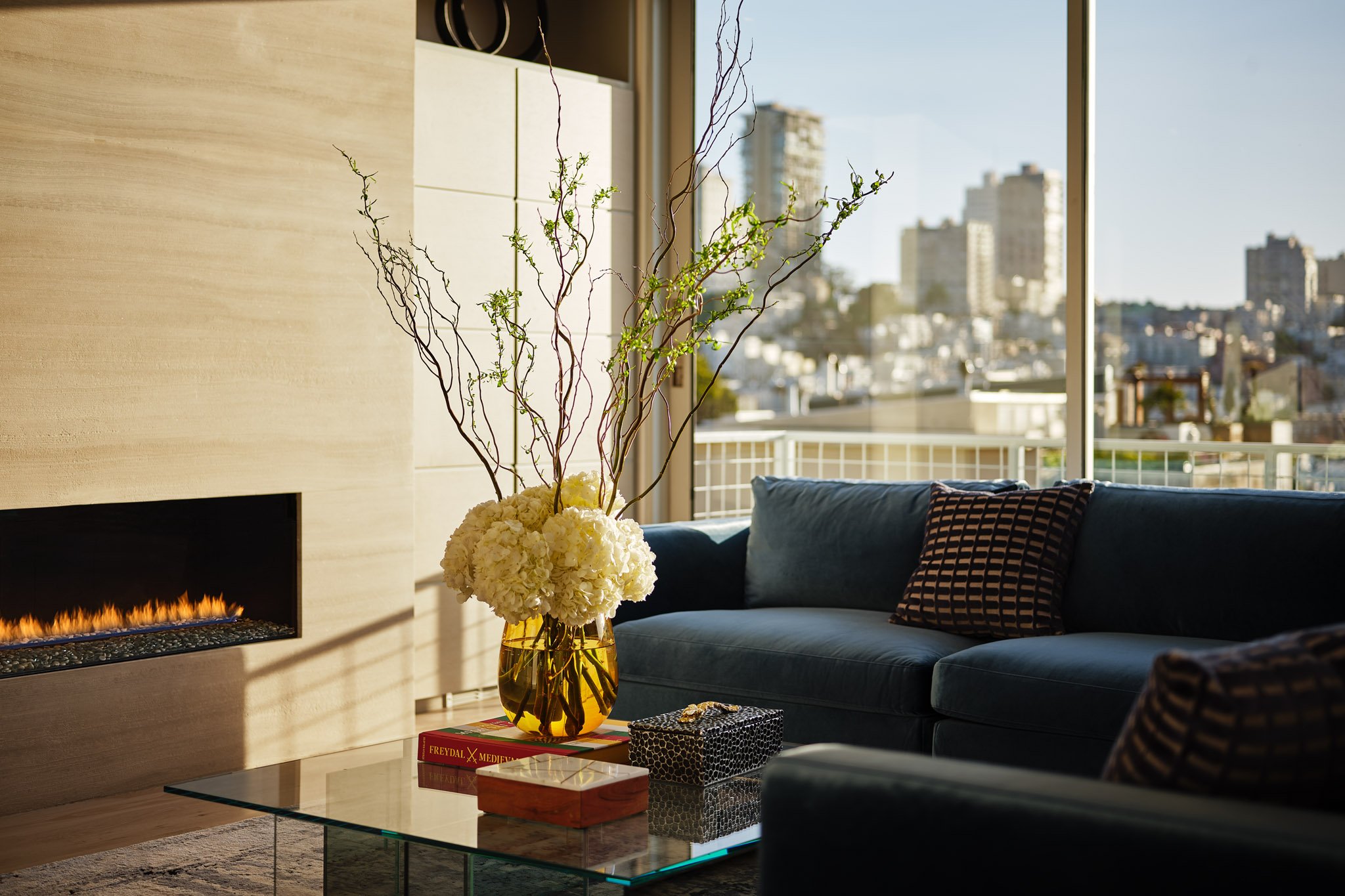 Modern living room with blue sofa and patterned pillows, glass coffee table with books and decorative box, large floral arrangement with branches in a gold vase, fireplace, and city view through large window.