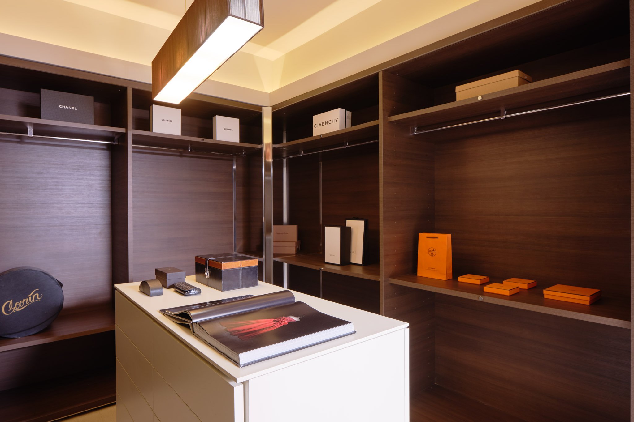 Empty walk-in closet with dark wood shelves, orange boxes on right shelf, white desk with open magazine, black box, and remote control, and a black round item labeled 'Gown' on lower left shelf.