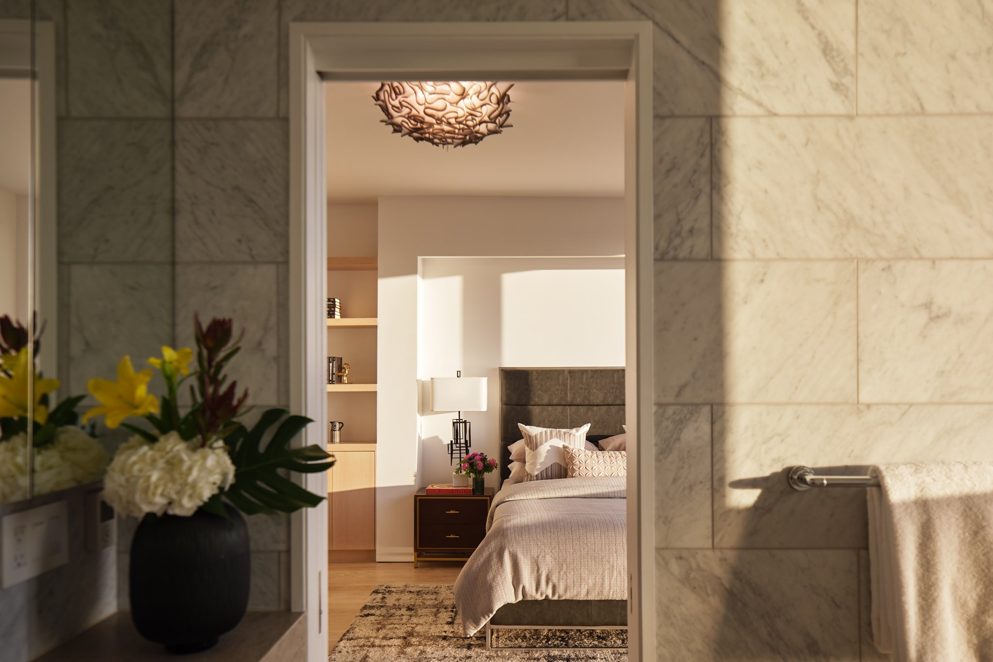 View of a bedroom through a doorway, with a bed, nightstand, lamp, and decorative pillows, seen from a bathroom with tiled walls and a vase of flowers.