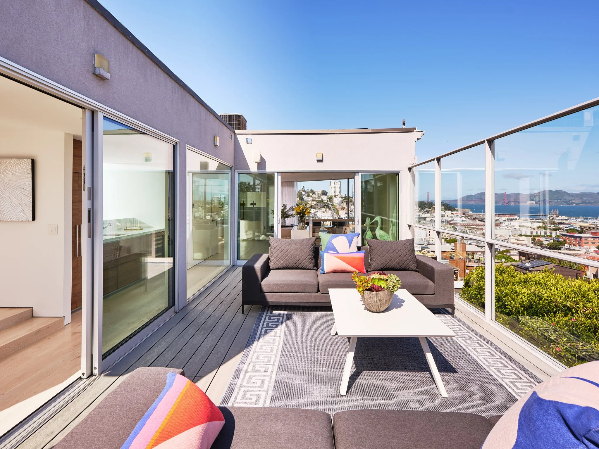 Modern rooftop balcony with outdoor furniture, glass railing, city view, and a bridge in the distance under a sunny sky.