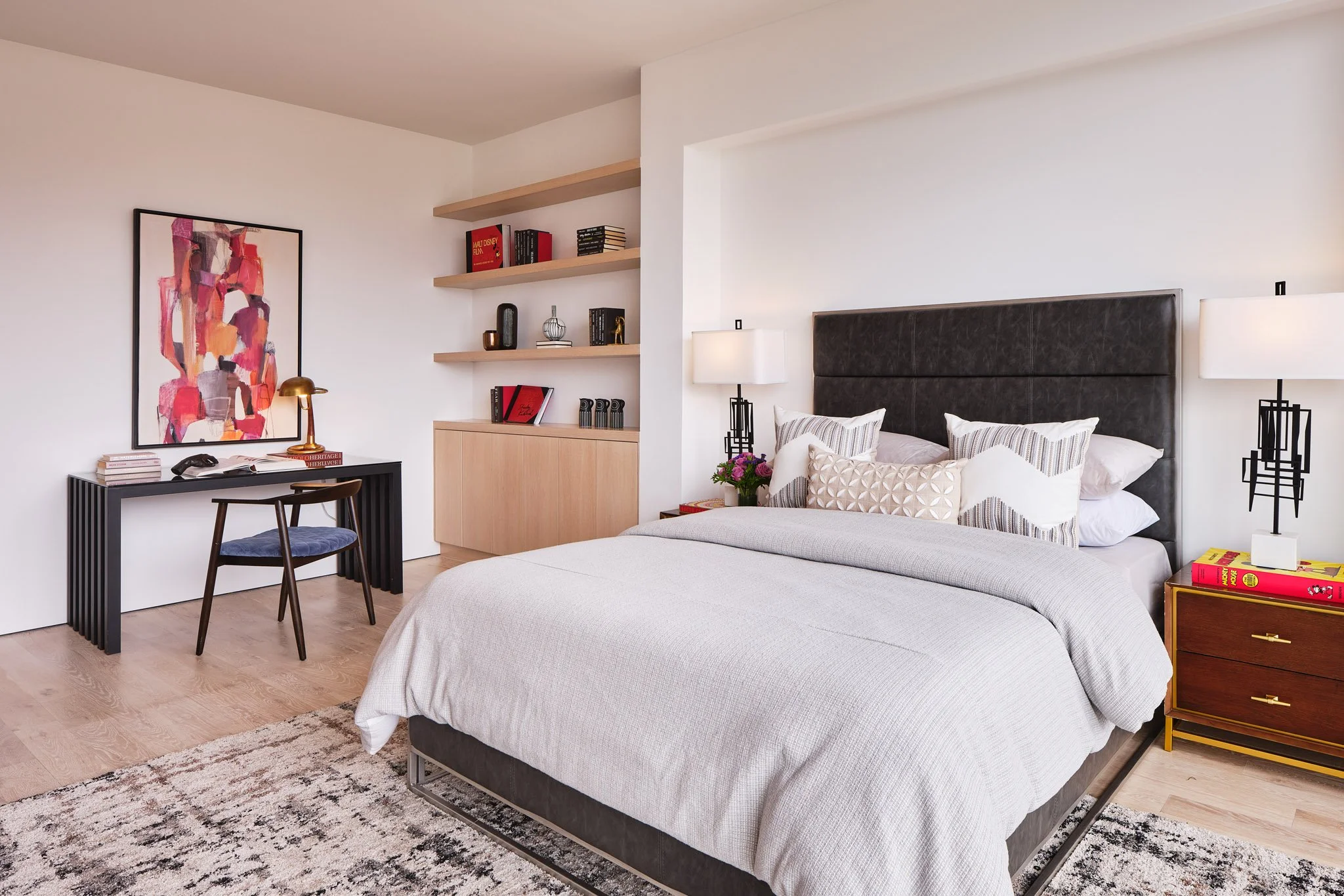 Modern bedroom with a bed, multiple pillows, and a black headboard. Nightstand with books and a lamp, a wall-mounted lamp, and a vase of flowers. Desk with a chair, table lamp, and framed abstract art on the wall. Shelves with books and decorative it