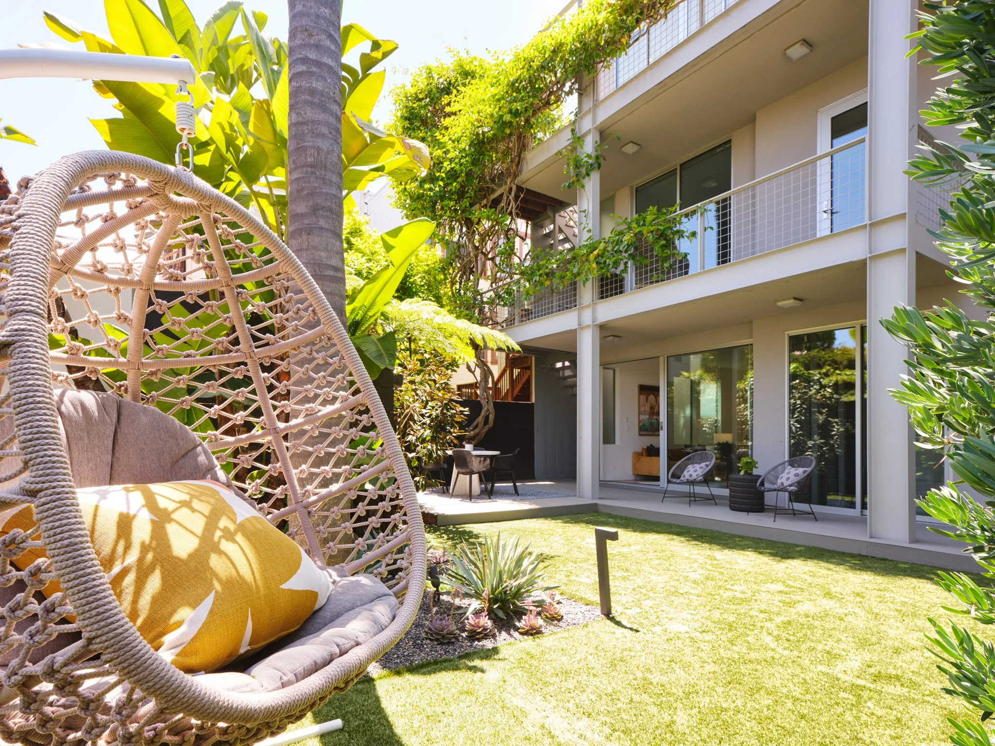 Modern apartment balcony with white railing, outdoor furniture, potted plants, and lush greenery in a sunny courtyard garden.