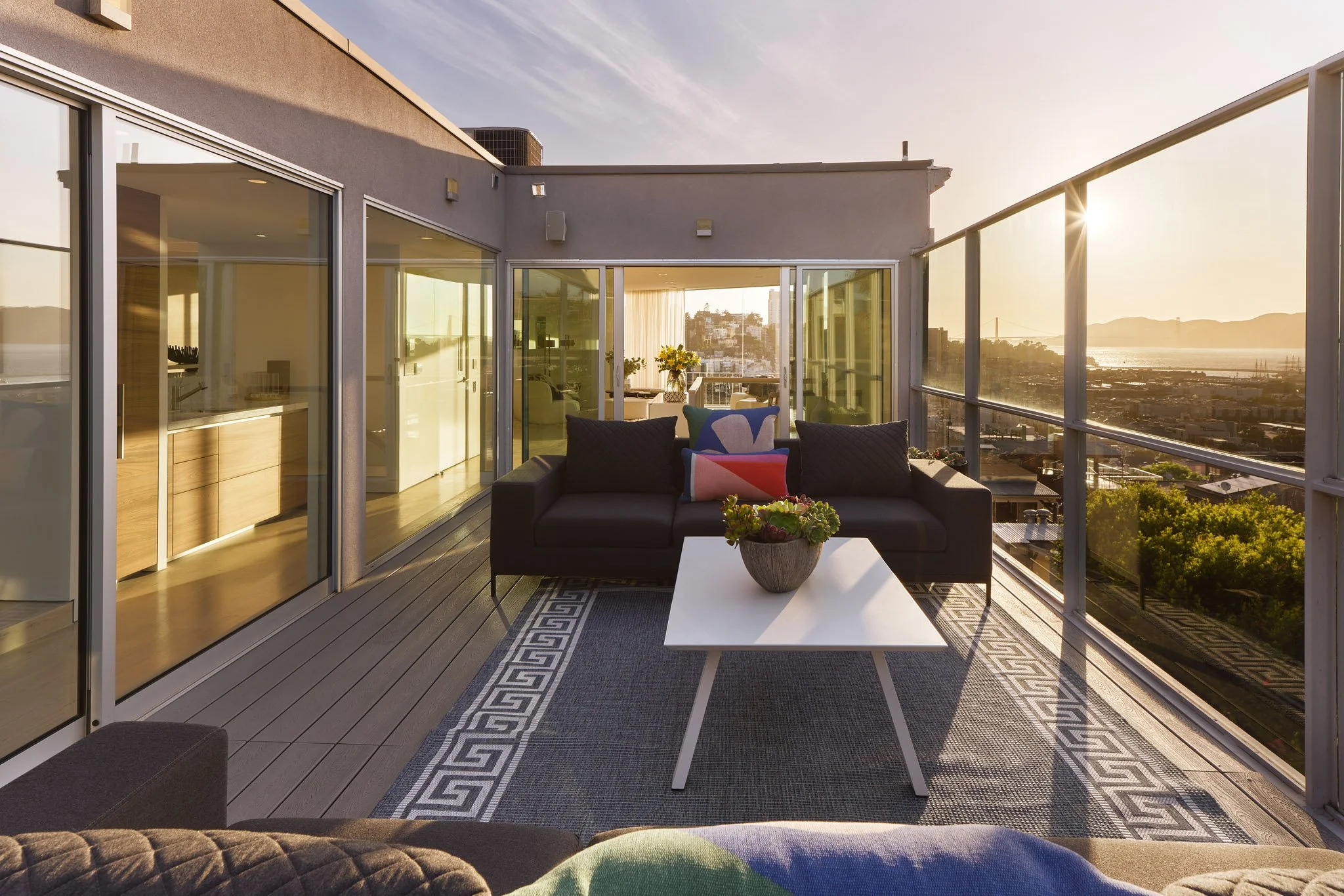 An outdoor balcony with glass railings, a black sofa with colorful pillows, a white coffee table with a potted plant, and a city skyline view during sunset.