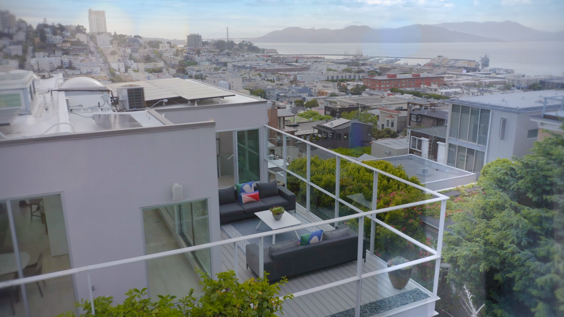 Outdoor balcony with black sofas, colorful pillows, white coffee table, and glass railing overlooking a cityscape and water with hills in the background.