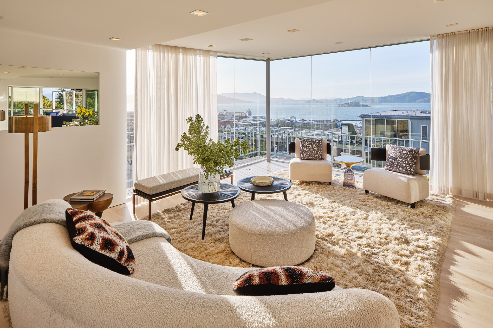 Modern living room with large glass windows showing a coastal view, featuring white furniture, a shaggy rug, potted plant, and decorative pillows.