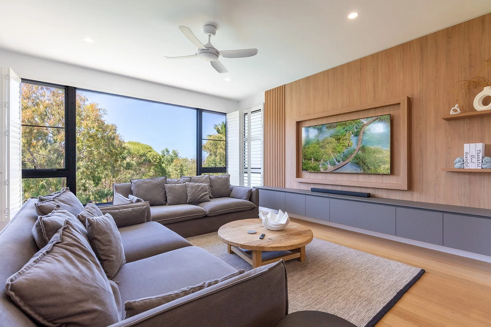 Modern living room with large gray sofas, round wooden coffee table, wall-mounted TV, and floor-to-ceiling windows.