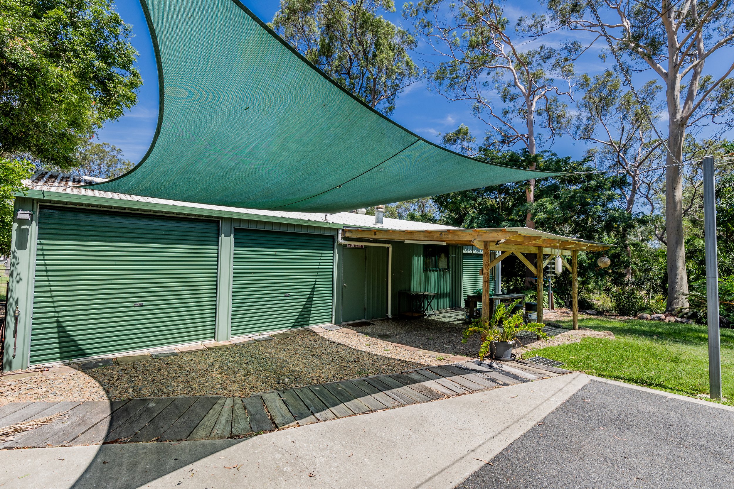 Green garage with metal doors under a shade sail, surrounded by trees and grass in a yard setting.